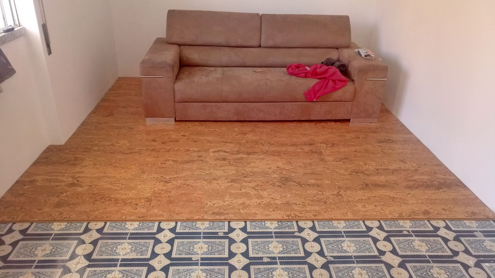 Sofá marrom sobre piso de cortiça elevado em uma sala com azulejos estampados. Roupas vermelhas estão sobre o sofá.