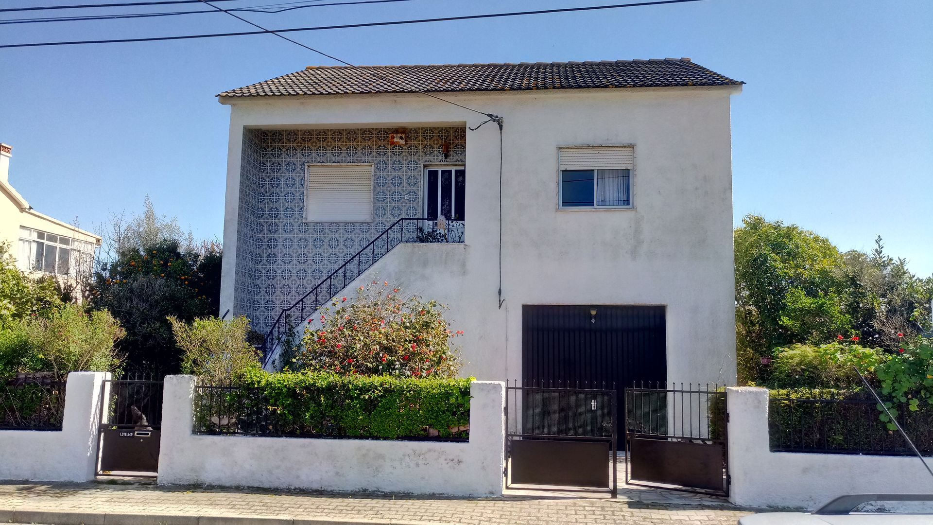 Casa branca de dois andares com portão e porta de garagem pretos. Arbustos e folhagens verdes cercam a propriedade.
