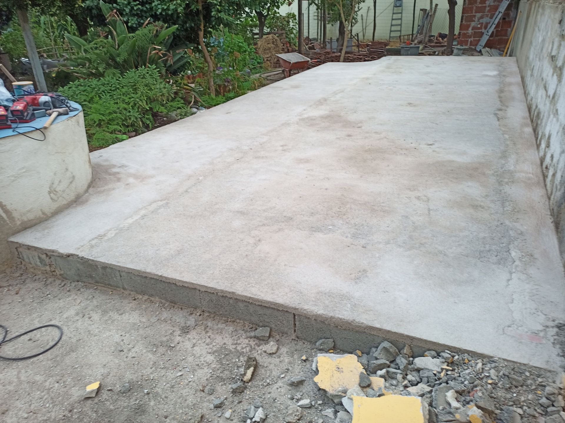 Laje de concreto em um quintal, delimitada por um muro baixo. Ferramentas e entulhos visíveis.