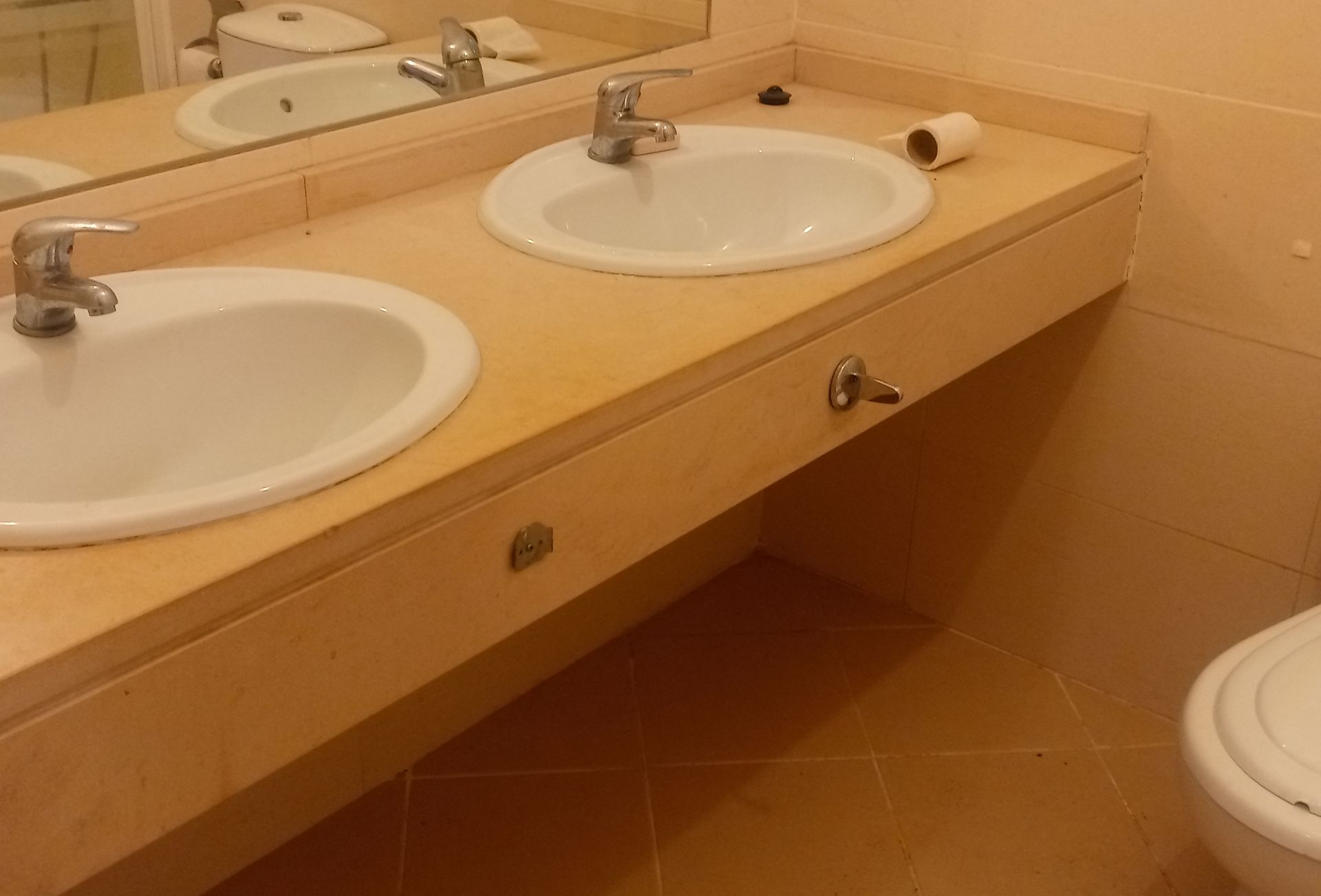 Banheiro com duas pias em uma bancada de cor clara, com um espelho acima e um vaso sanitário ao lado.