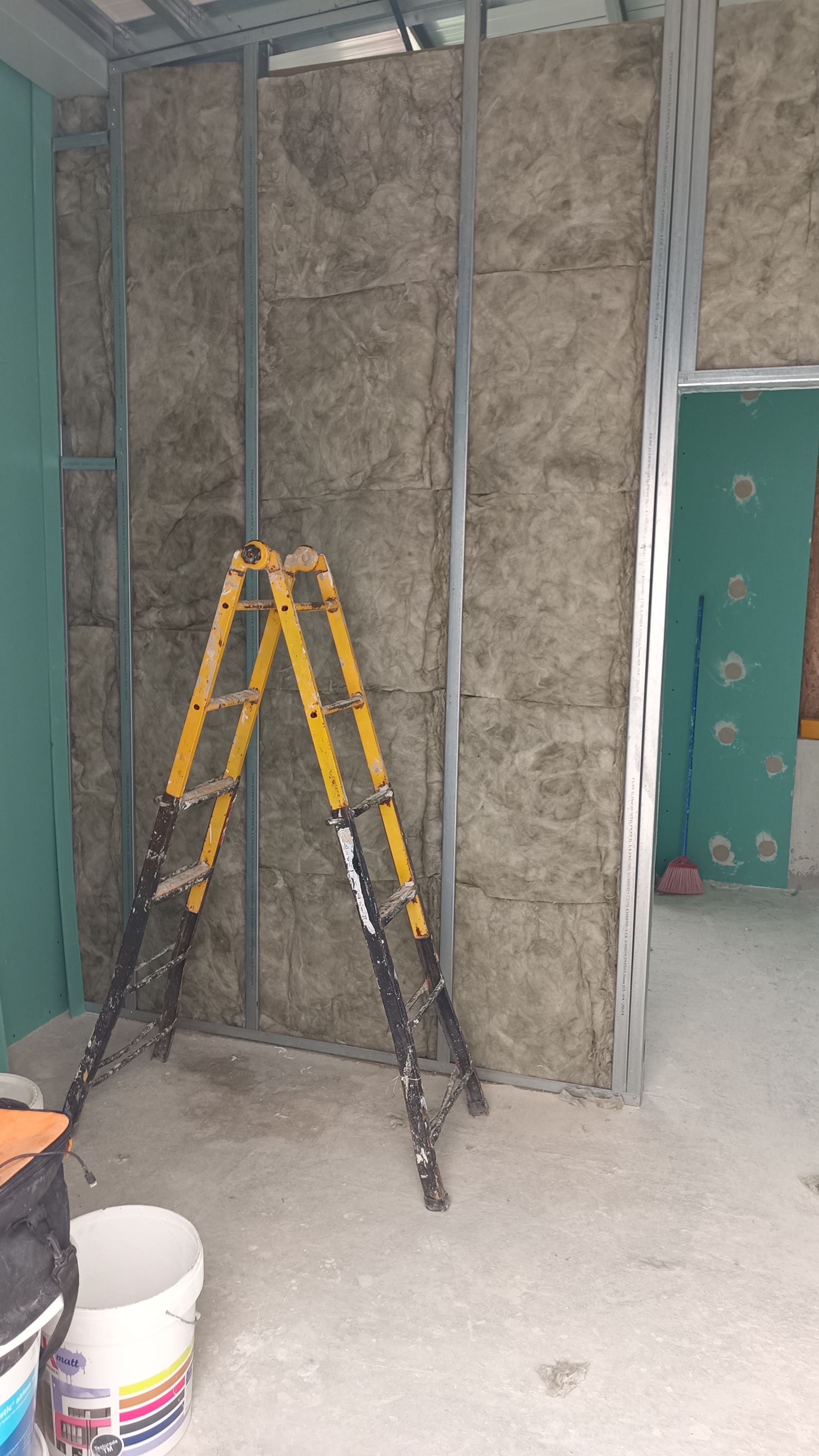 Construção da parede interna: perfis metálicos, isolamento e uma escada amarela em um cômodo.