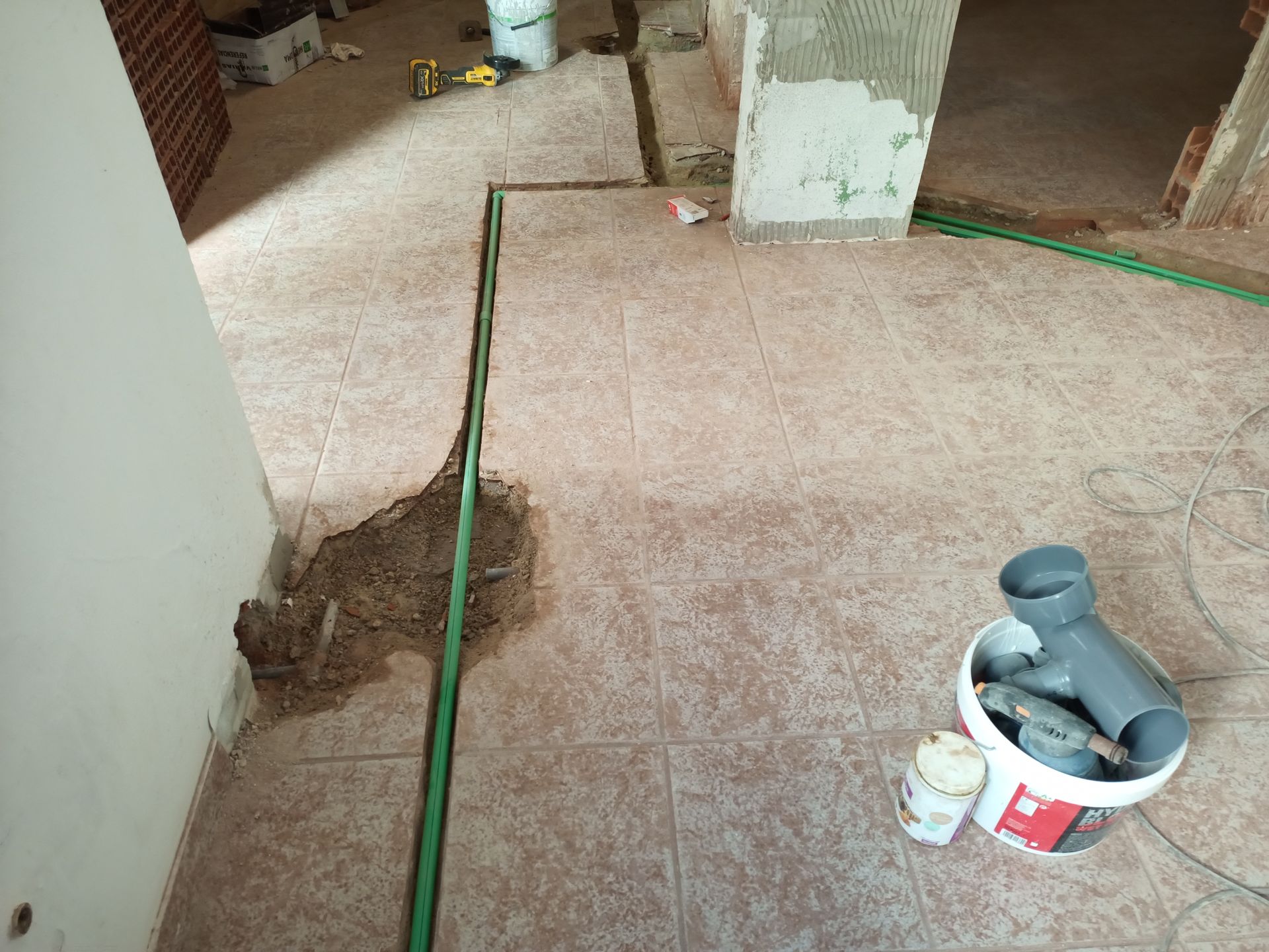Reforma em andamento: cano verde instalado em uma vala no piso de azulejos. Baldes com materiais hidráulicos à direita.
