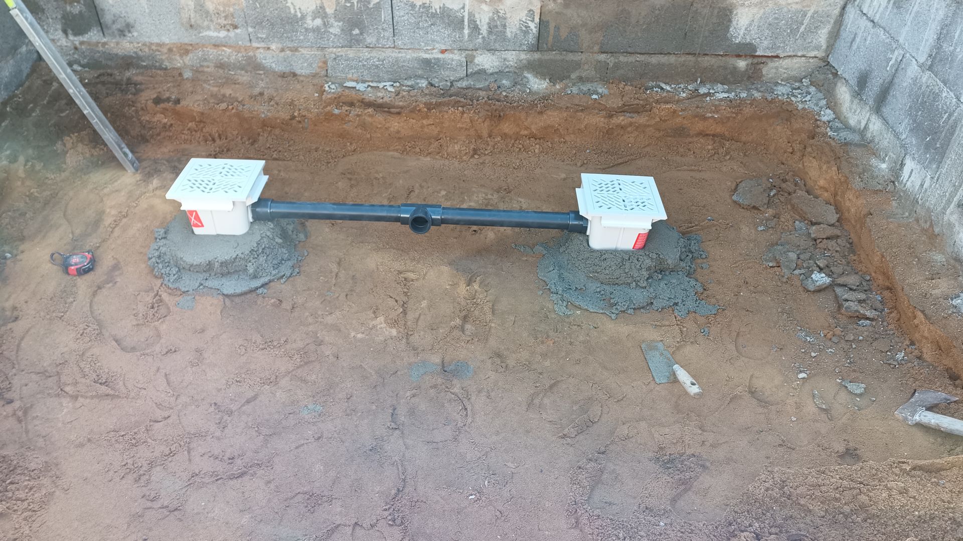 Duas caixas de drenagem conectadas por um tubo, instaladas em cascalho dentro de uma área cercada por paredes de concreto.