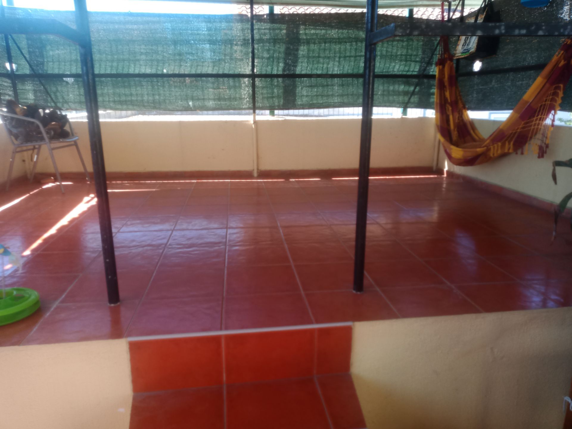 Terraço com piso de ladrilhos vermelhos, uma rede e postes de sustentação de metal sob um telhado sombreado.