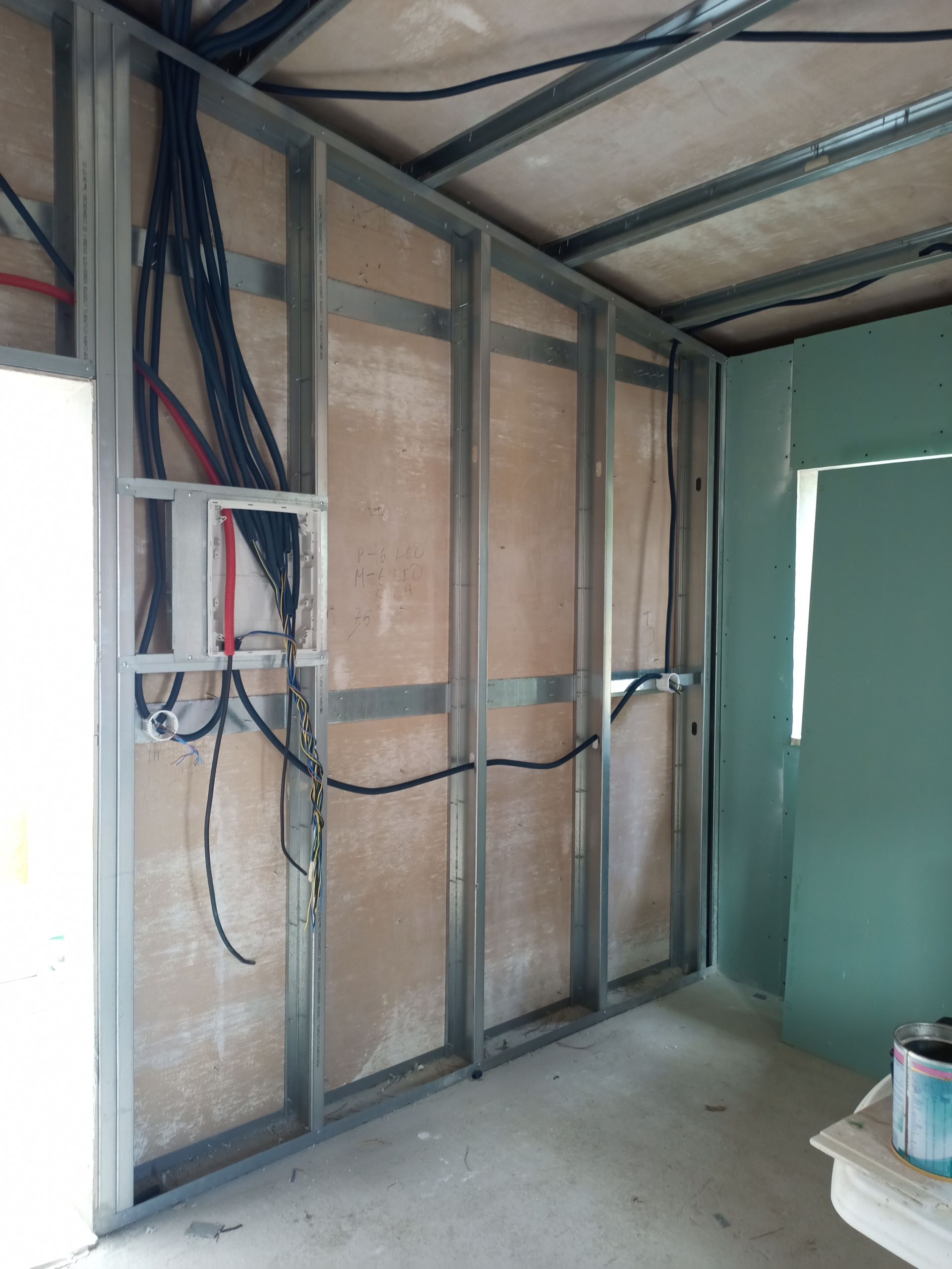 Parede interna em construção com estrutura metálica, fiação exposta e parte da parede pintada de verde.