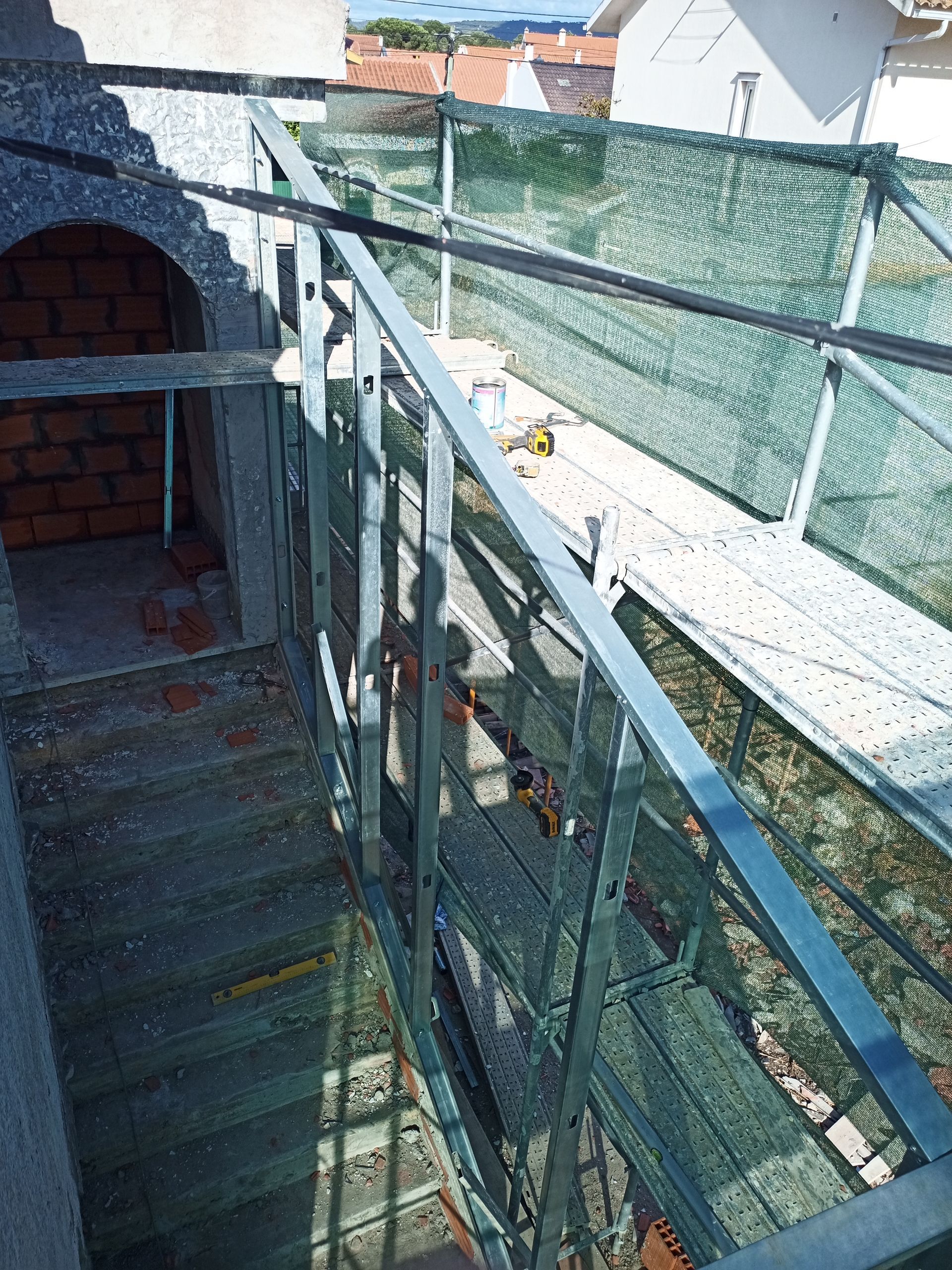 Corrimão de metal em passarela externa, em um canteiro de obras. Rede verde e prédio de tijolos também são visíveis.