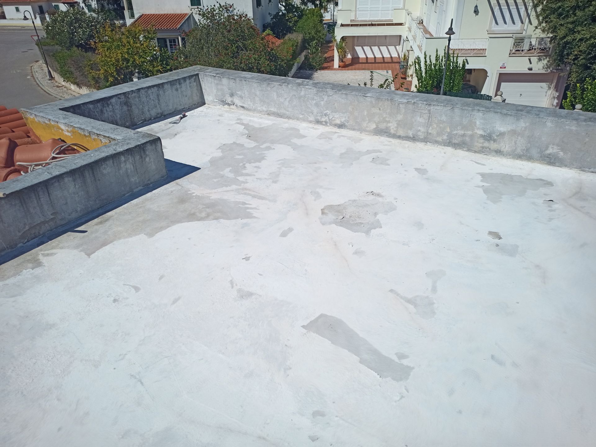Telhado plano com revestimento branco, borda de concreto, casas e árvores ao fundo.