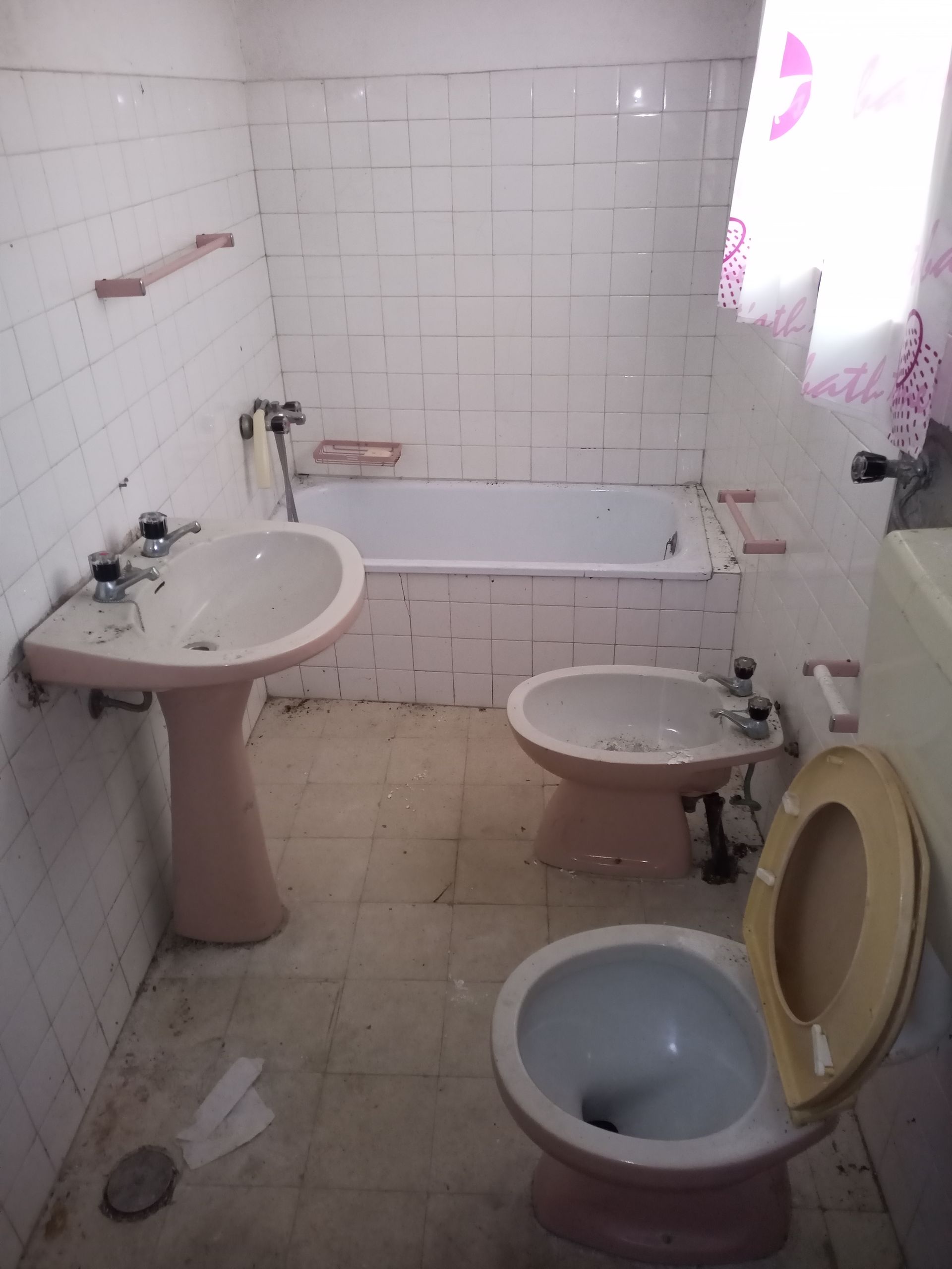 Banheiro rosa com pia, bidê, vaso sanitário e banheira; paredes e piso revestidos com azulejos, acessórios antigos.