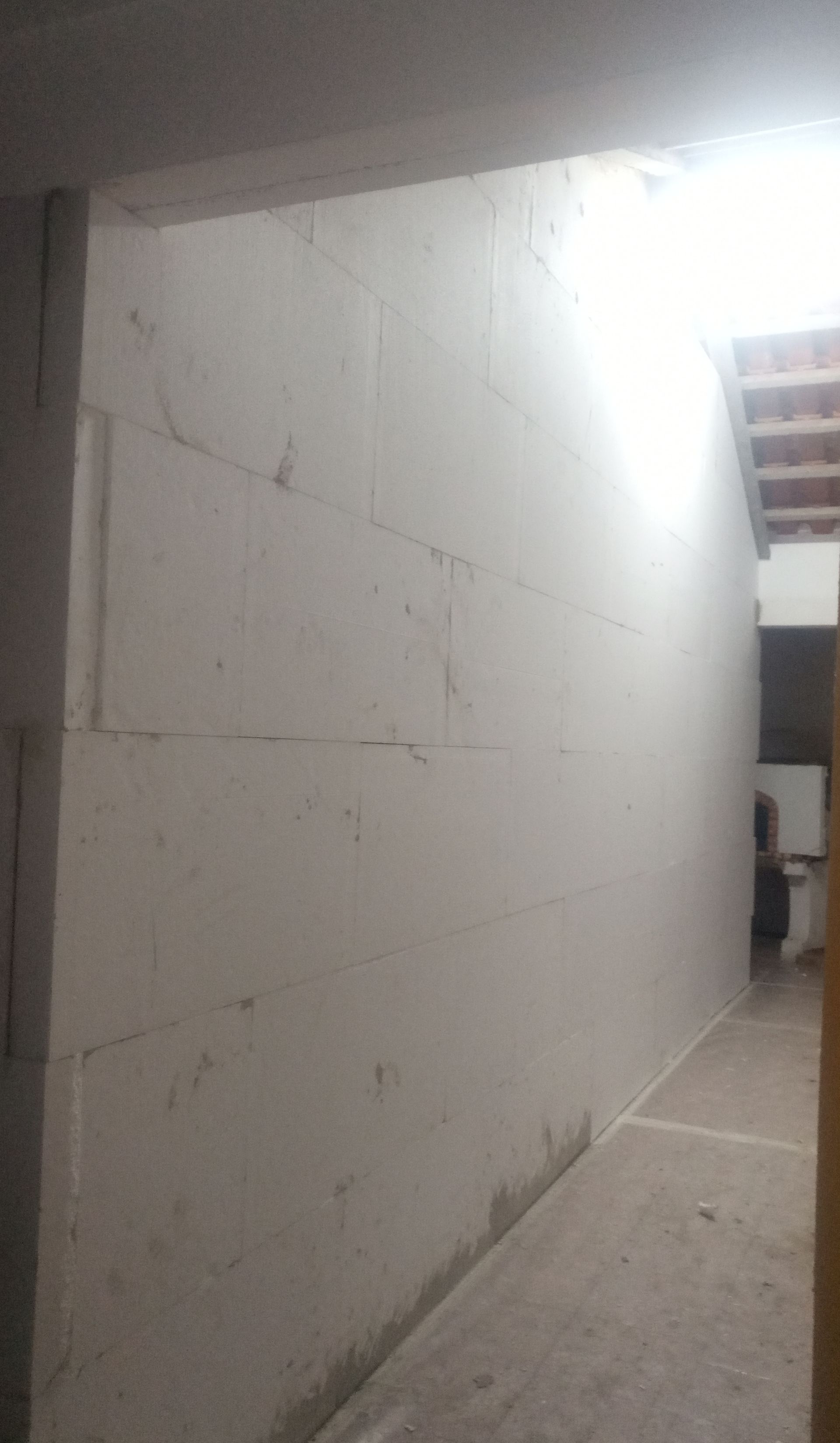 Paredes de blocos de concreto branco em um corredor, iluminadas por cima; piso de concreto; porta no final.