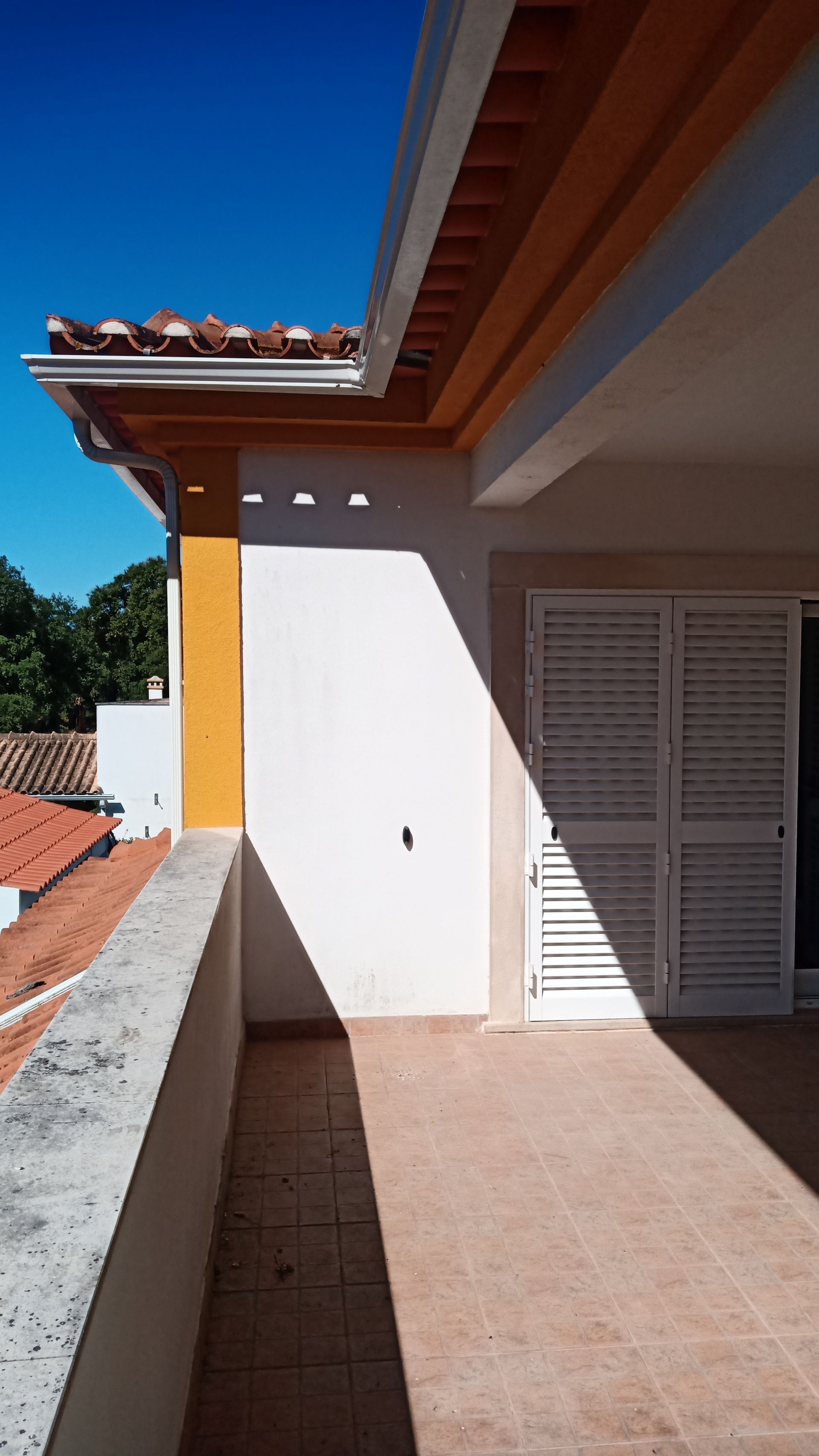 Varanda com longa sombra projetada pelo telhado, céu azul claro, telhas de terracota, parede branca e porta veneziana.