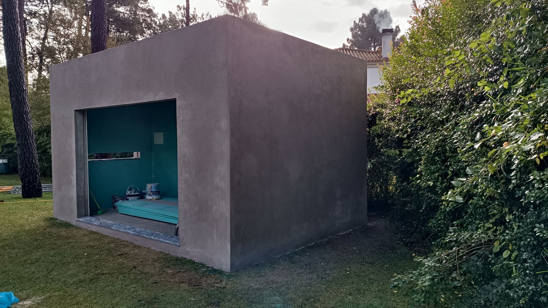 Estrutura em forma de caixa de concreto cinza, com entrada aberta, rodeada por folhagem verde, em uma área gramada com árvores.