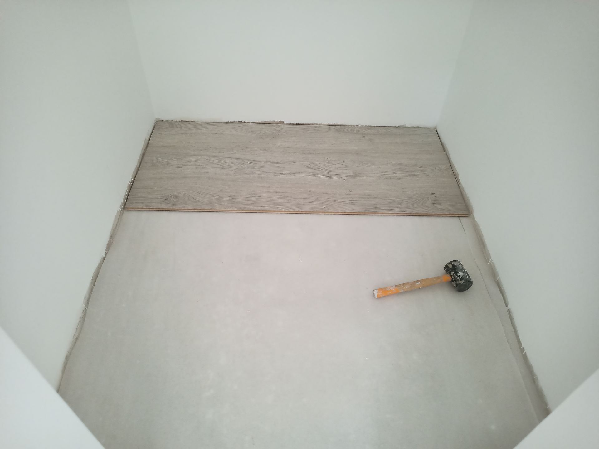 Sala interna com piso inacabado, parcialmente revestido com azulejos. Uma marreta está apoiada no chão.