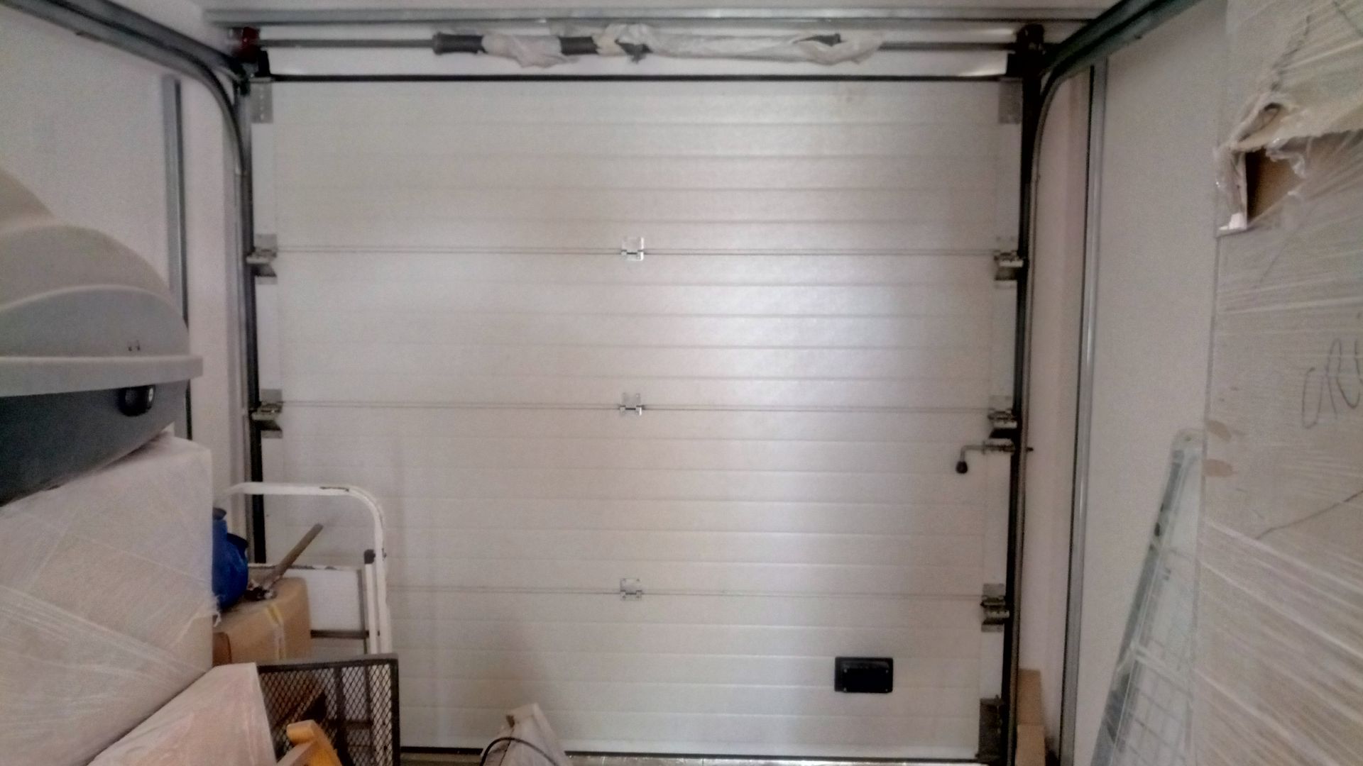 Porta de garagem branca, fechada, com trilhos e ferragens de metal. Vista interna.