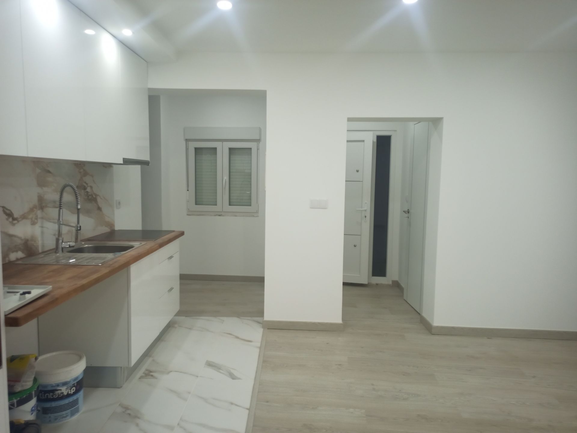 Vista interna de uma cozinha e cômodos adjacentes. Apresenta armários brancos, bancada de madeira e backsplash de mármore.