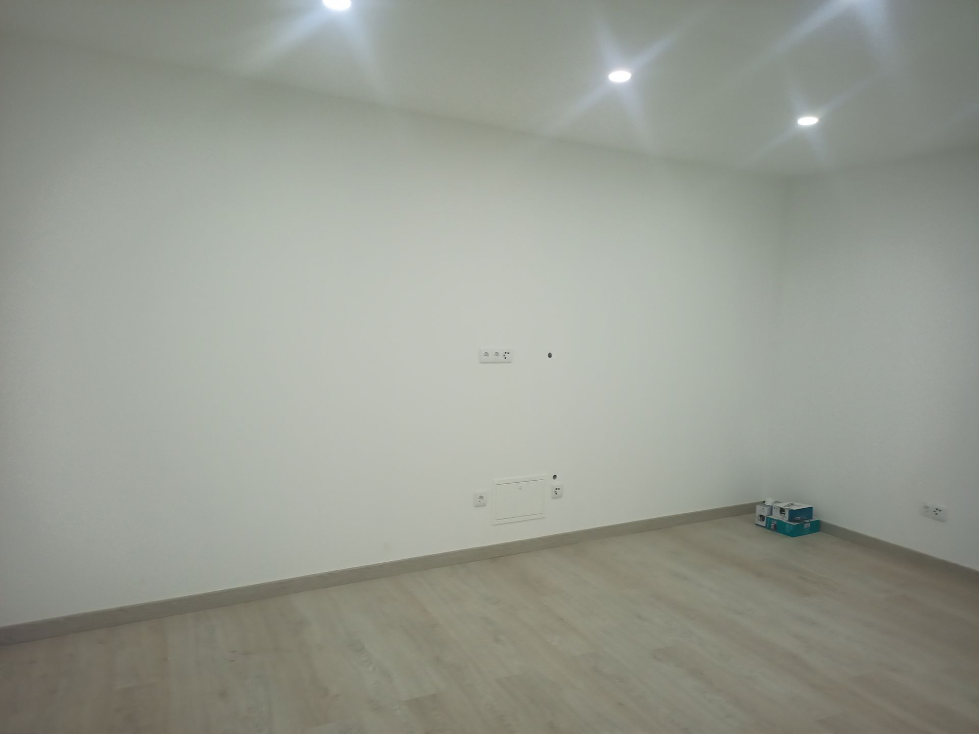 Sala vazia com paredes brancas, piso de madeira clara e luzes embutidas no teto.