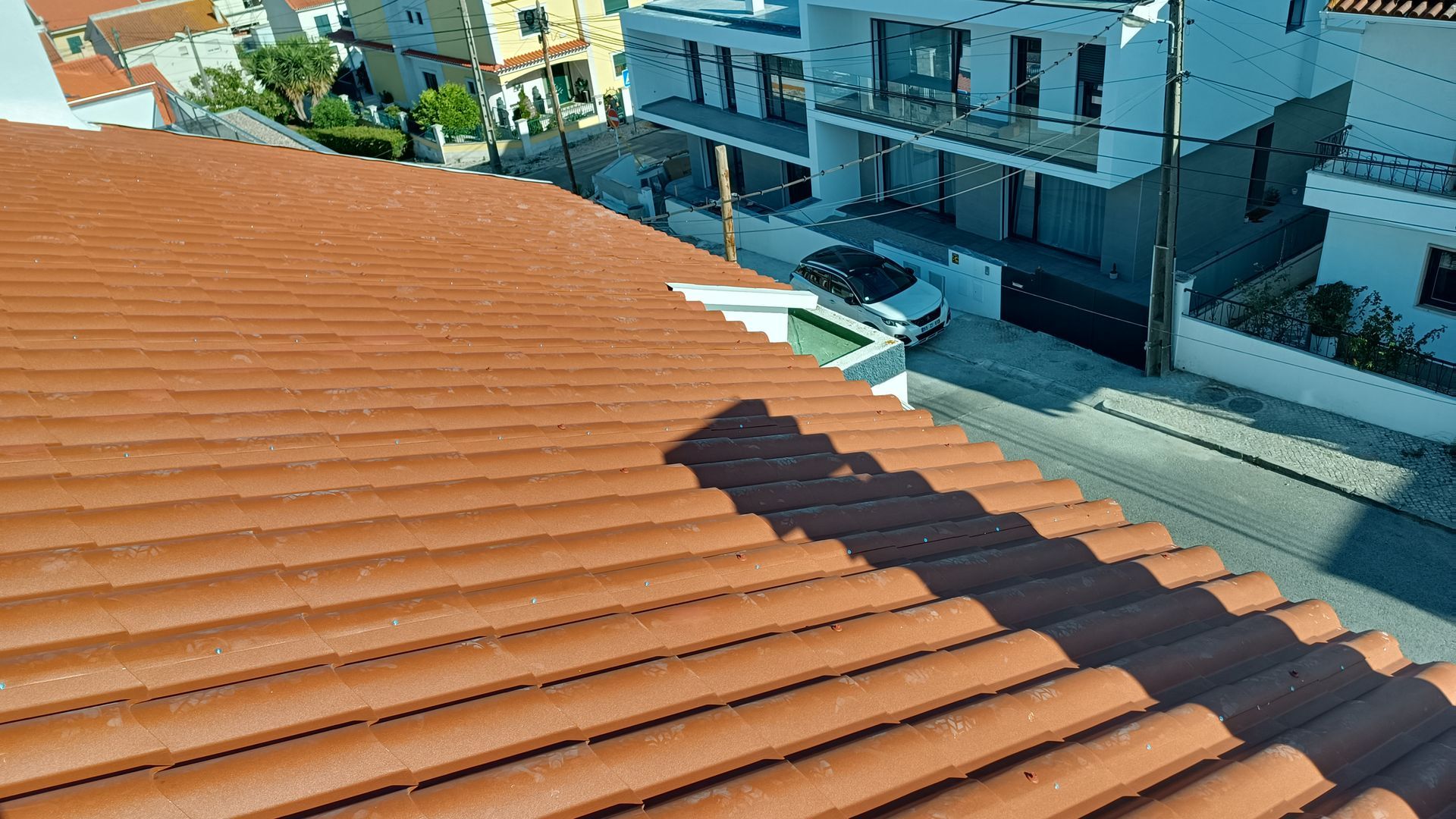 Telhado de telhas de terracota projetando sombra sobre uma rua com prédios e um carro estacionado.
