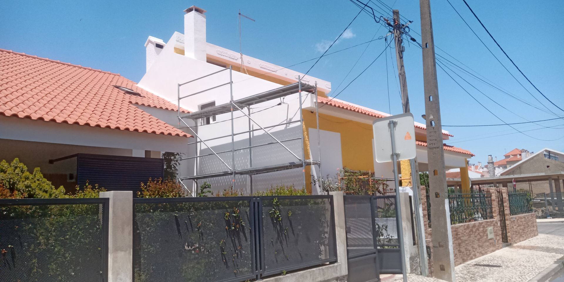 Uma fileira de casas com telhados de telha vermelha e paredes brancas. Em uma delas, há um andaime montado. Um poste de luz está na rua.