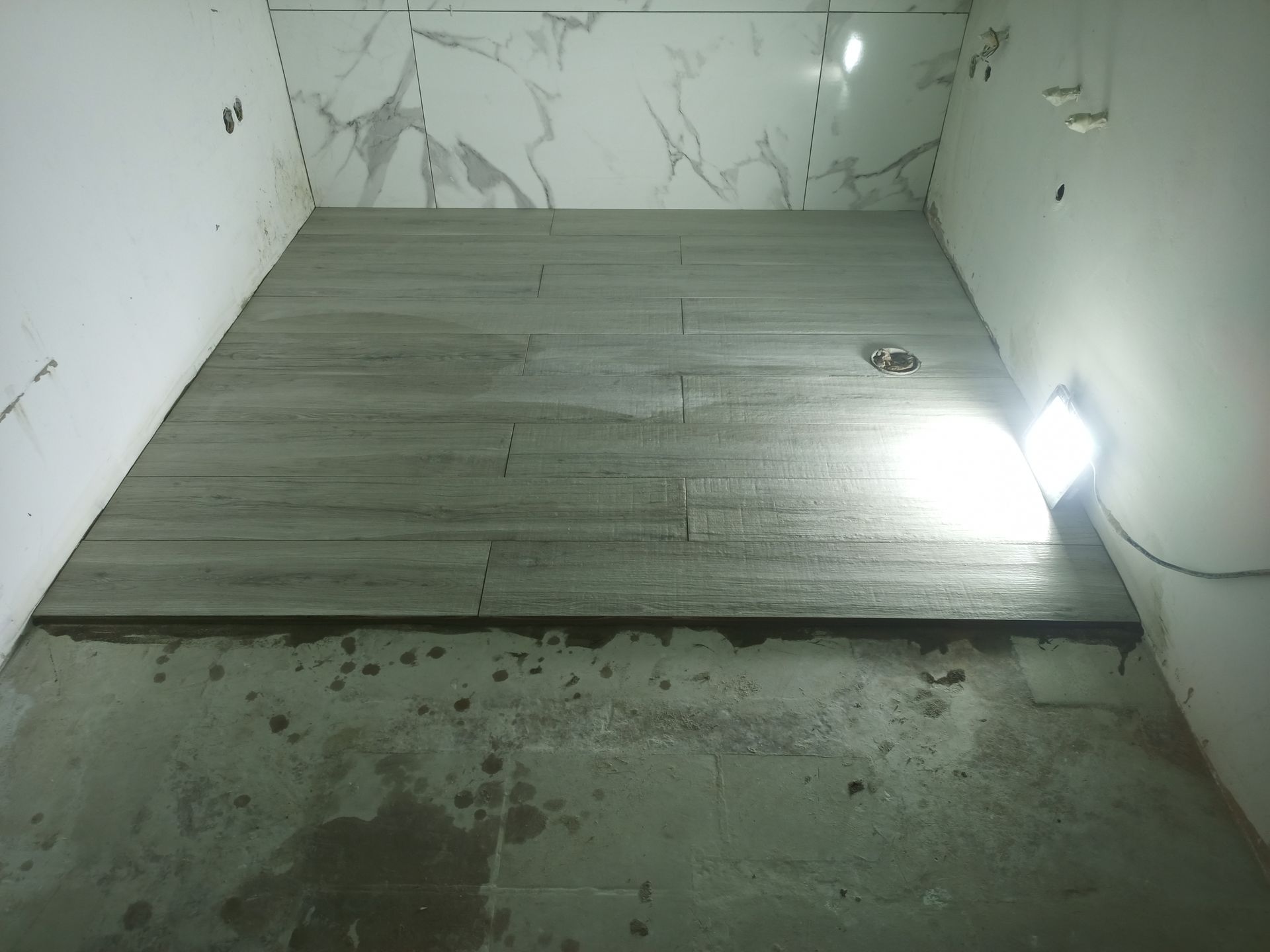 Banheiro em construção; piso de porcelanato imitando madeira instalado, azulejos brancos na parede. Iluminação de trabalho eficiente.