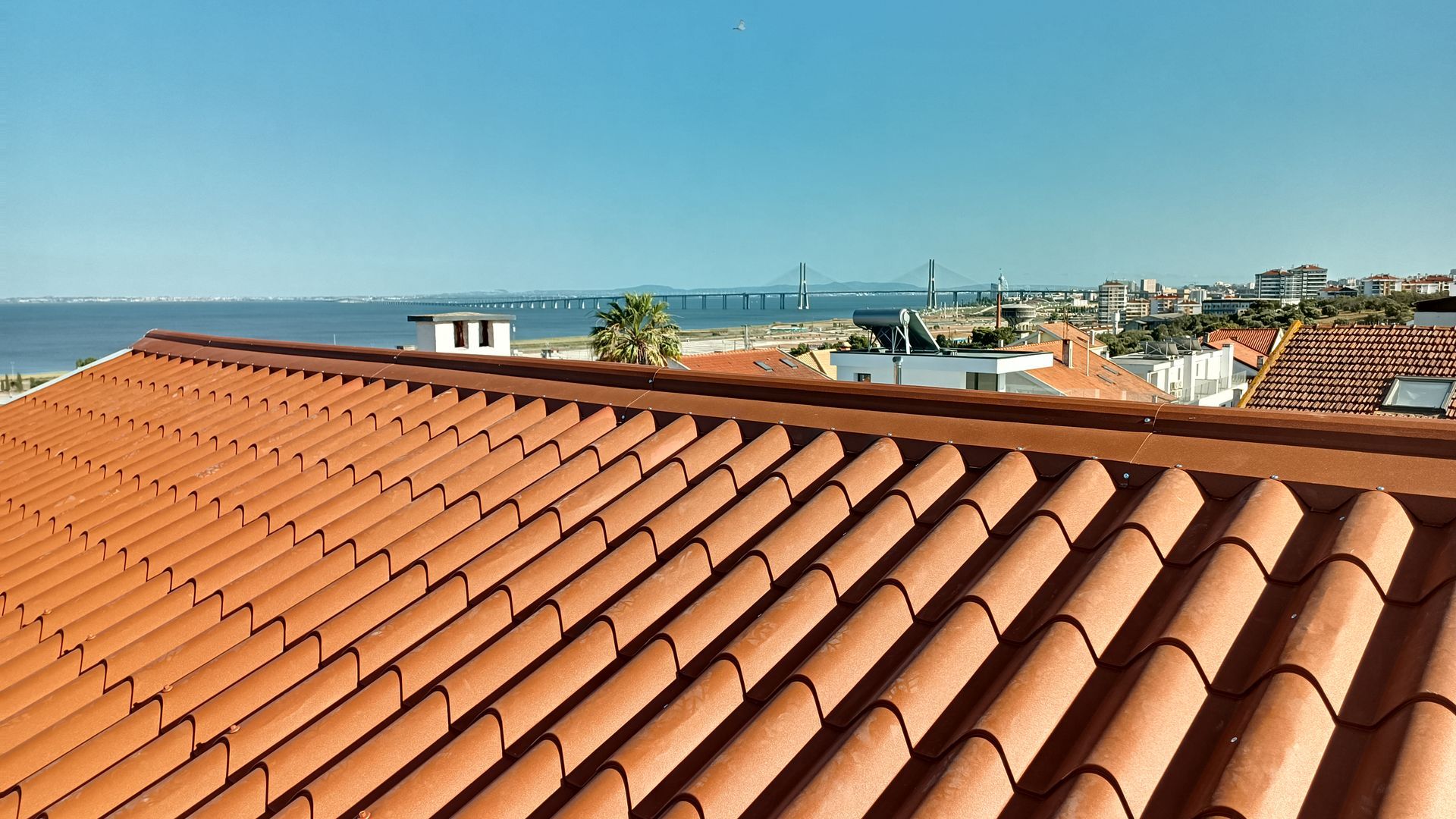 Telhado de telhas de terracota com vista para o oceano, céu azul e paisagem urbana ao longe.