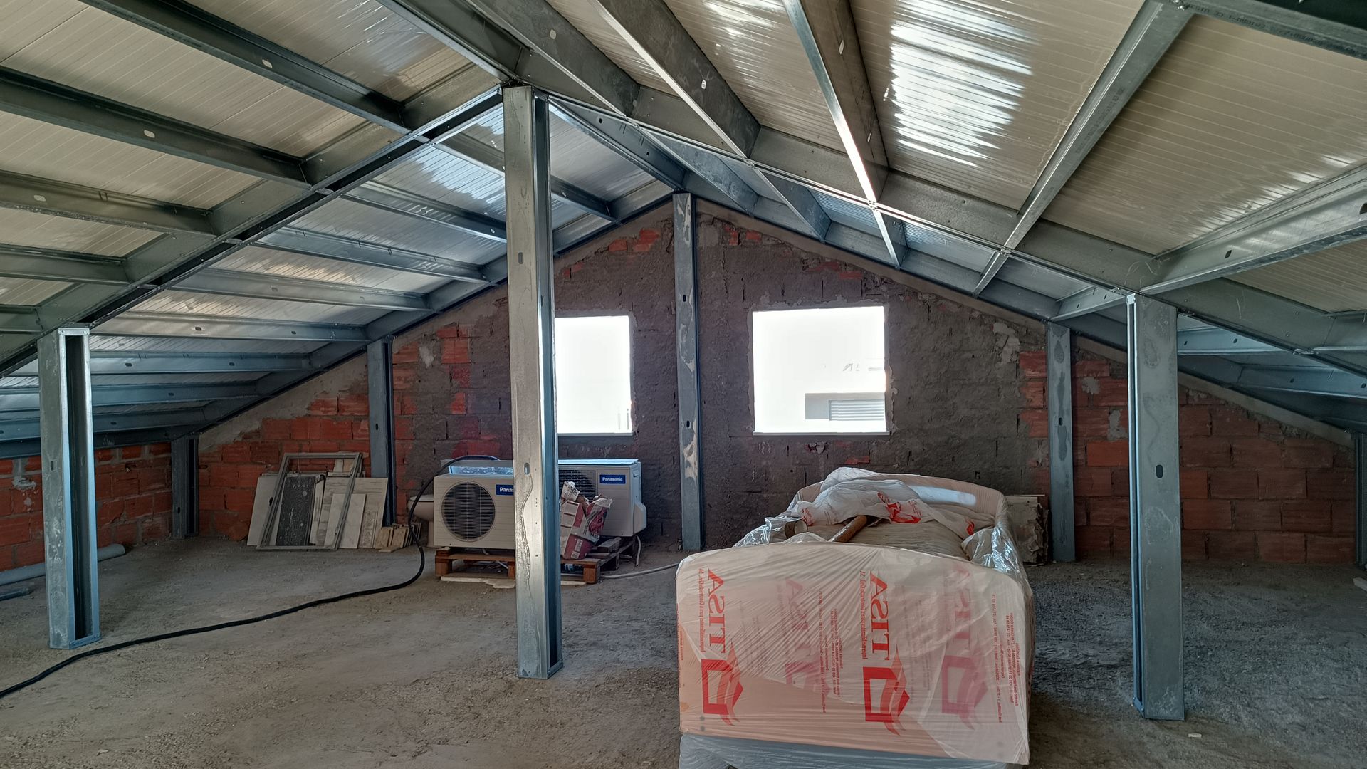 Interior de um sótão inacabado com vigas de aço expostas e paredes de tijolo. Duas janelas e materiais de construção são visíveis.