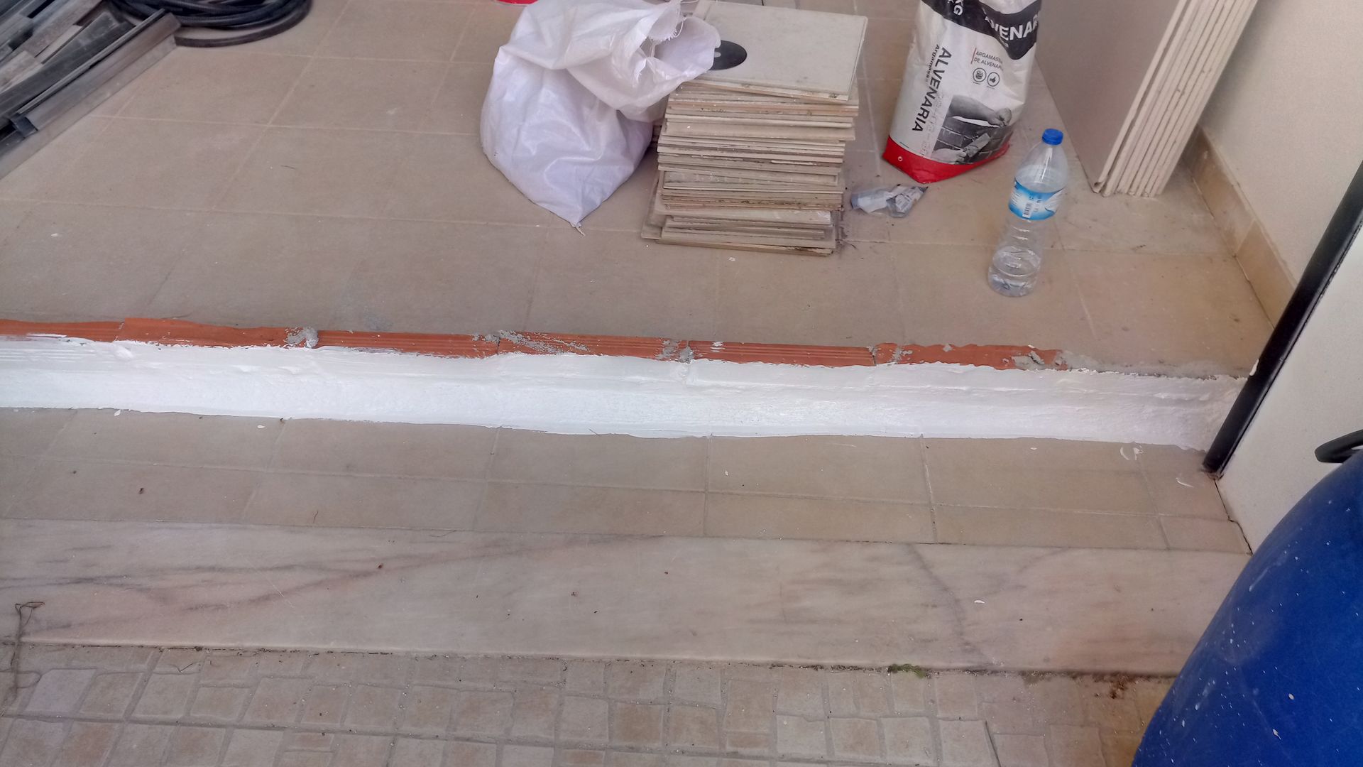 Construção em andamento. Piso de azulejo, rejunte branco, borda marrom. Sacolas, azulejos e uma garrafa de água estão espalhados pelo chão.