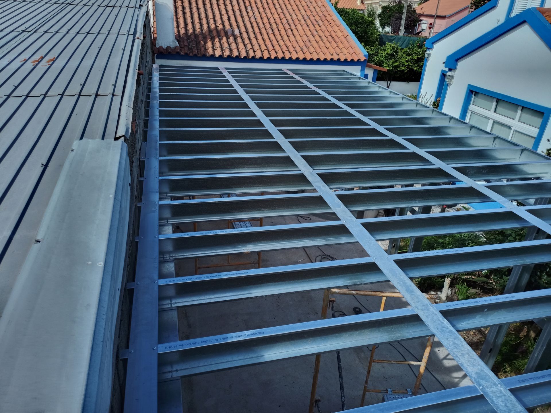 Estrutura metálica de um telhado em construção, em contraste com edifícios existentes com telhados azuis e vermelhos.