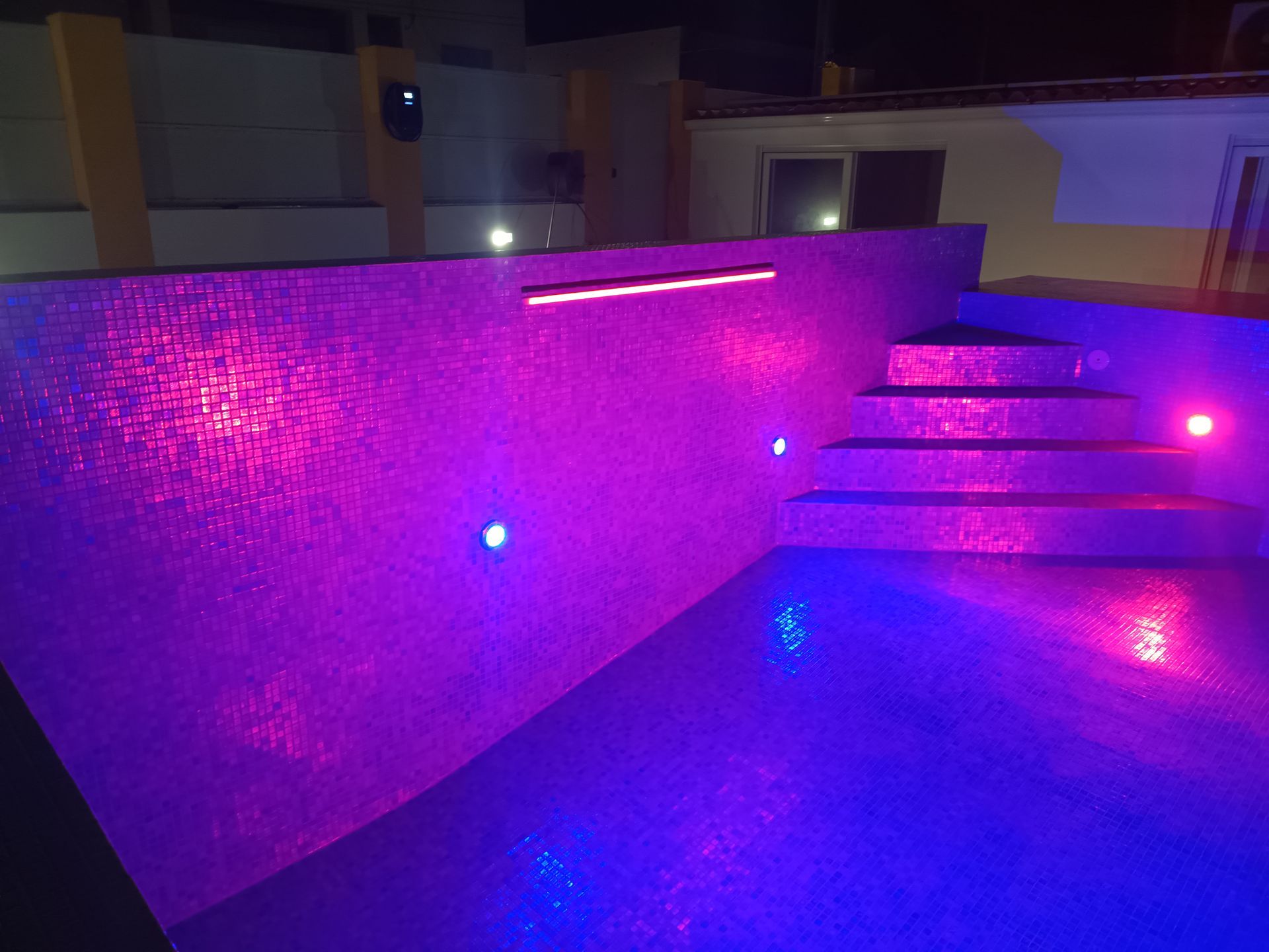 Piscina iluminada à noite com degraus e azulejos em mosaico, iluminada com luzes rosa e azuis.