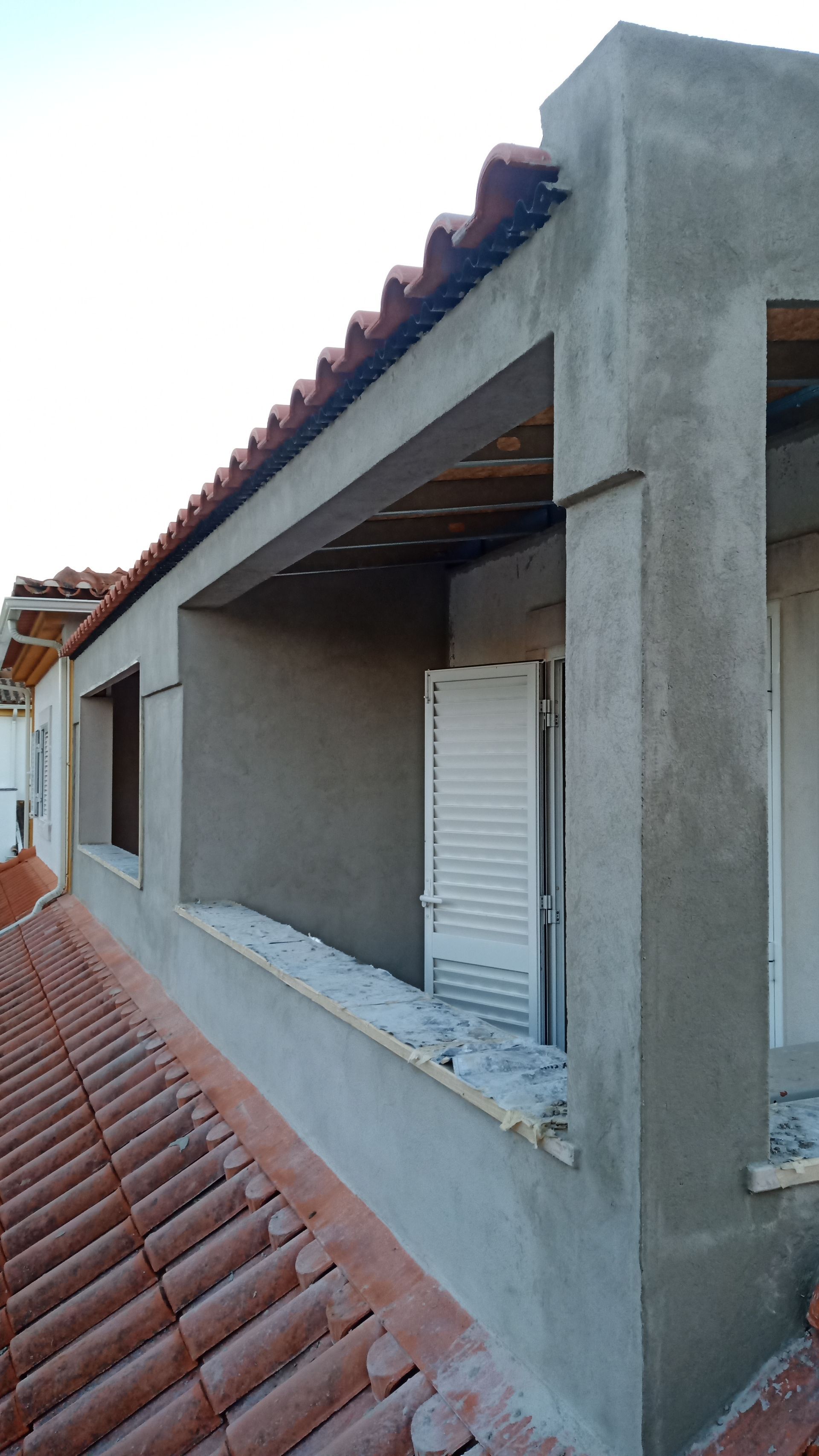 Vista do exterior de um edifício com telhado de telhas avermelhadas, paredes de estuque cinza e uma janela com venezianas brancas.