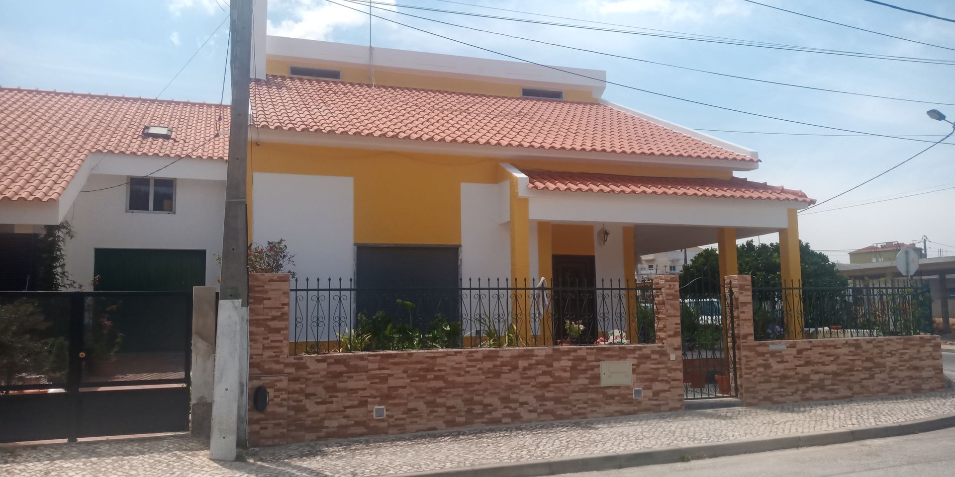 Casa amarela com telhado de telhas vermelhas, parede de tijolos e cerca de metal preta.