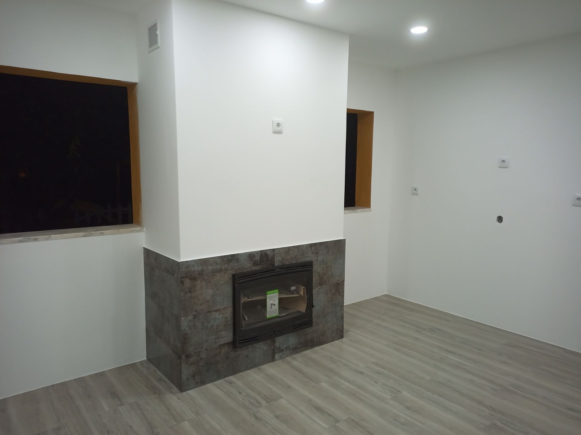 Sala vazia com lareira. Lareira de pedra cinza, paredes brancas, janelas com moldura de madeira, piso cinza claro.