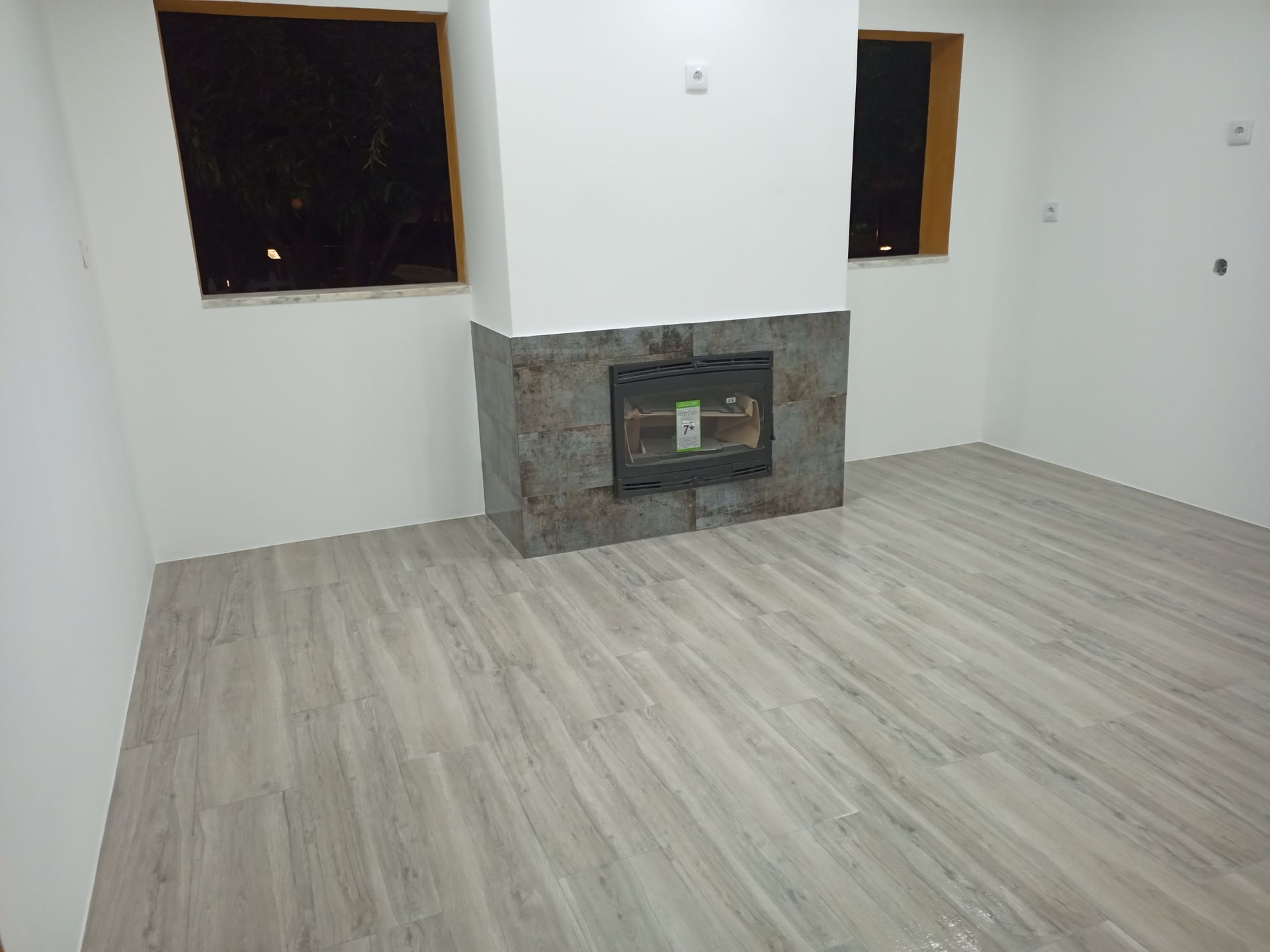 Sala vazia com lareira, piso de madeira e duas janelas. Paredes brancas, fachada da lareira em pedra.
