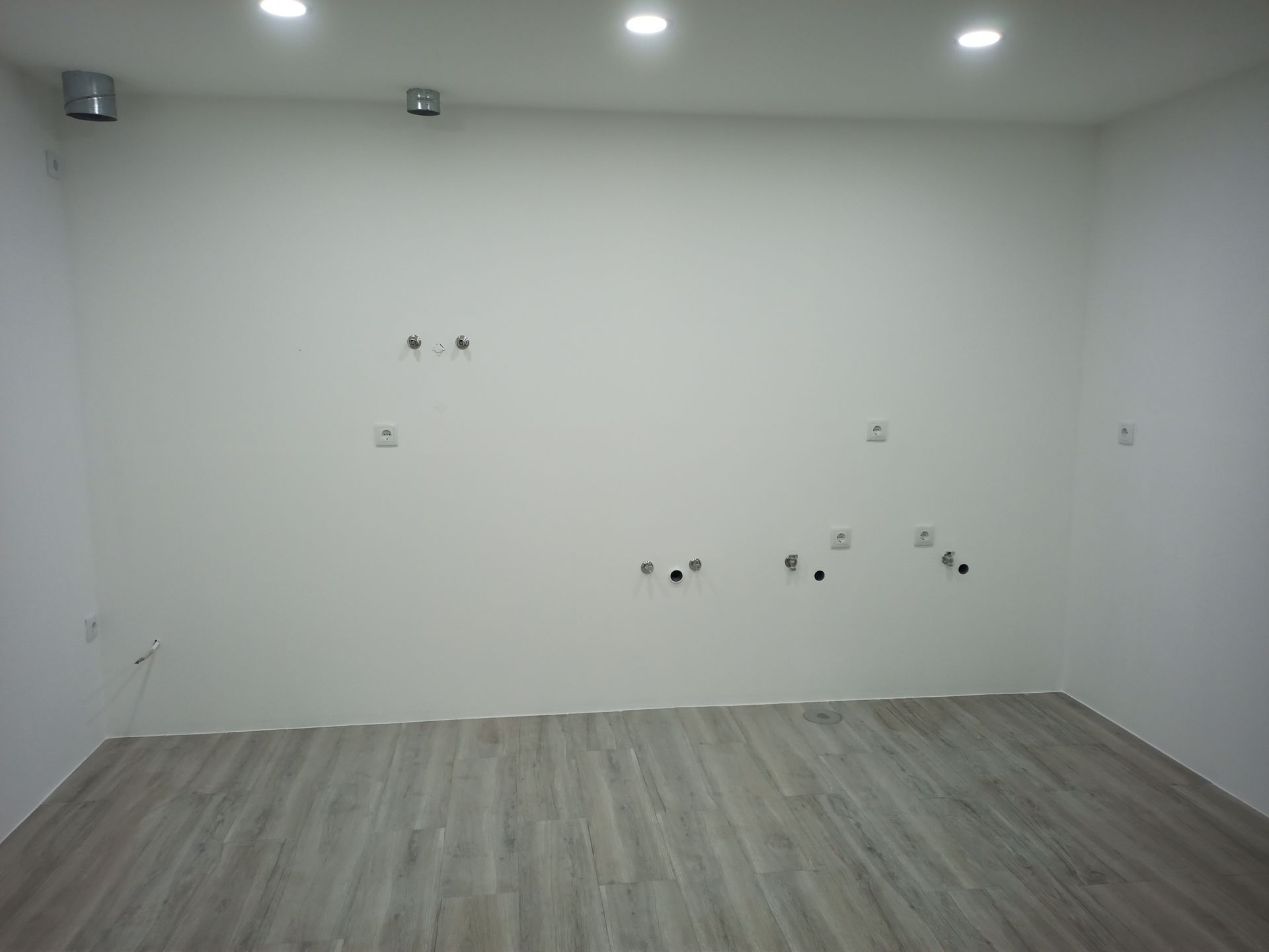 Sala vazia com piso de madeira cinza claro, paredes brancas e iluminação embutida. Diversos orifícios e acessórios são visíveis nas paredes.