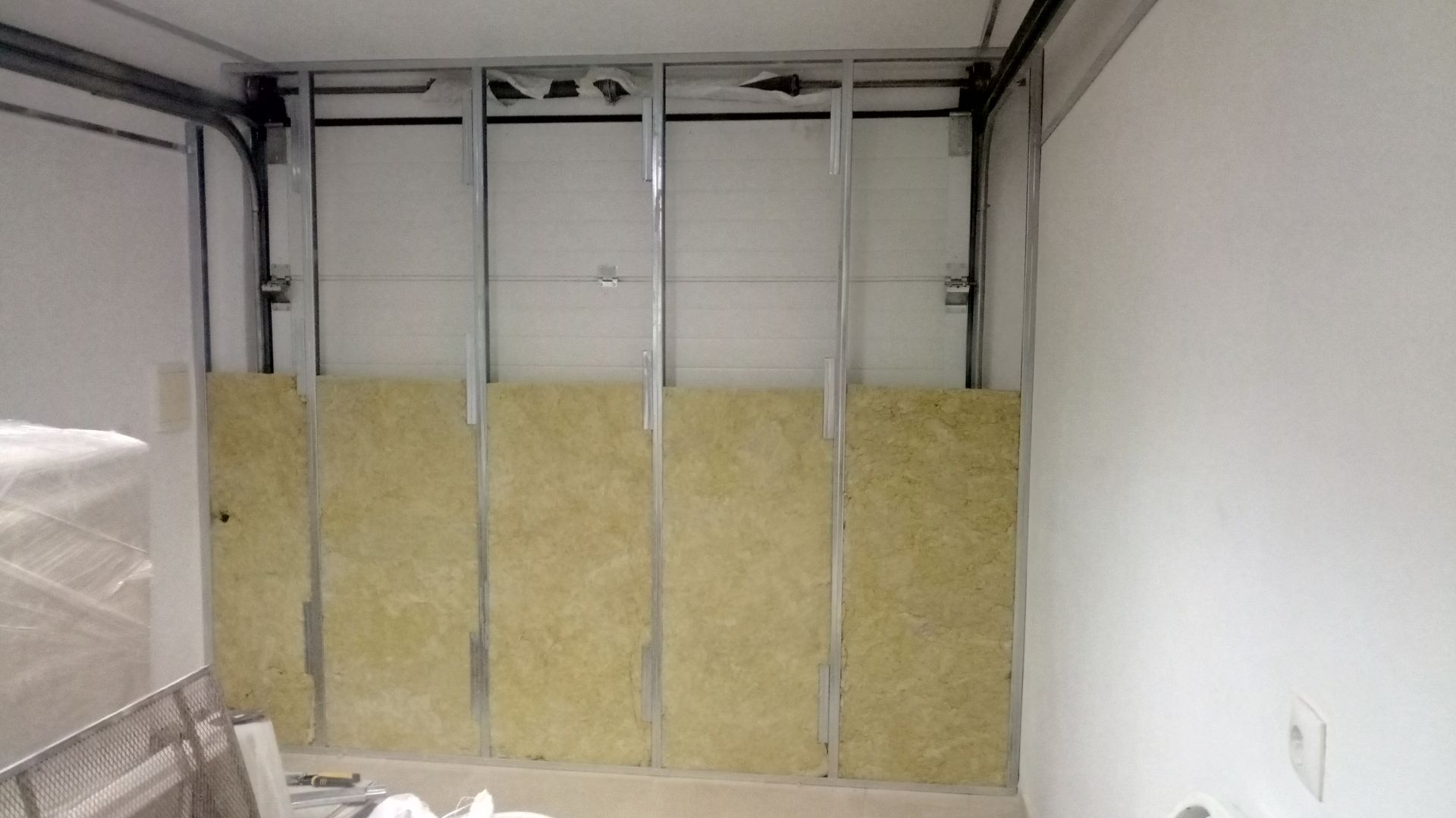 Uma parede interna parcialmente construída com perfis metálicos, isolamento e parede branca, provavelmente em uma garagem.