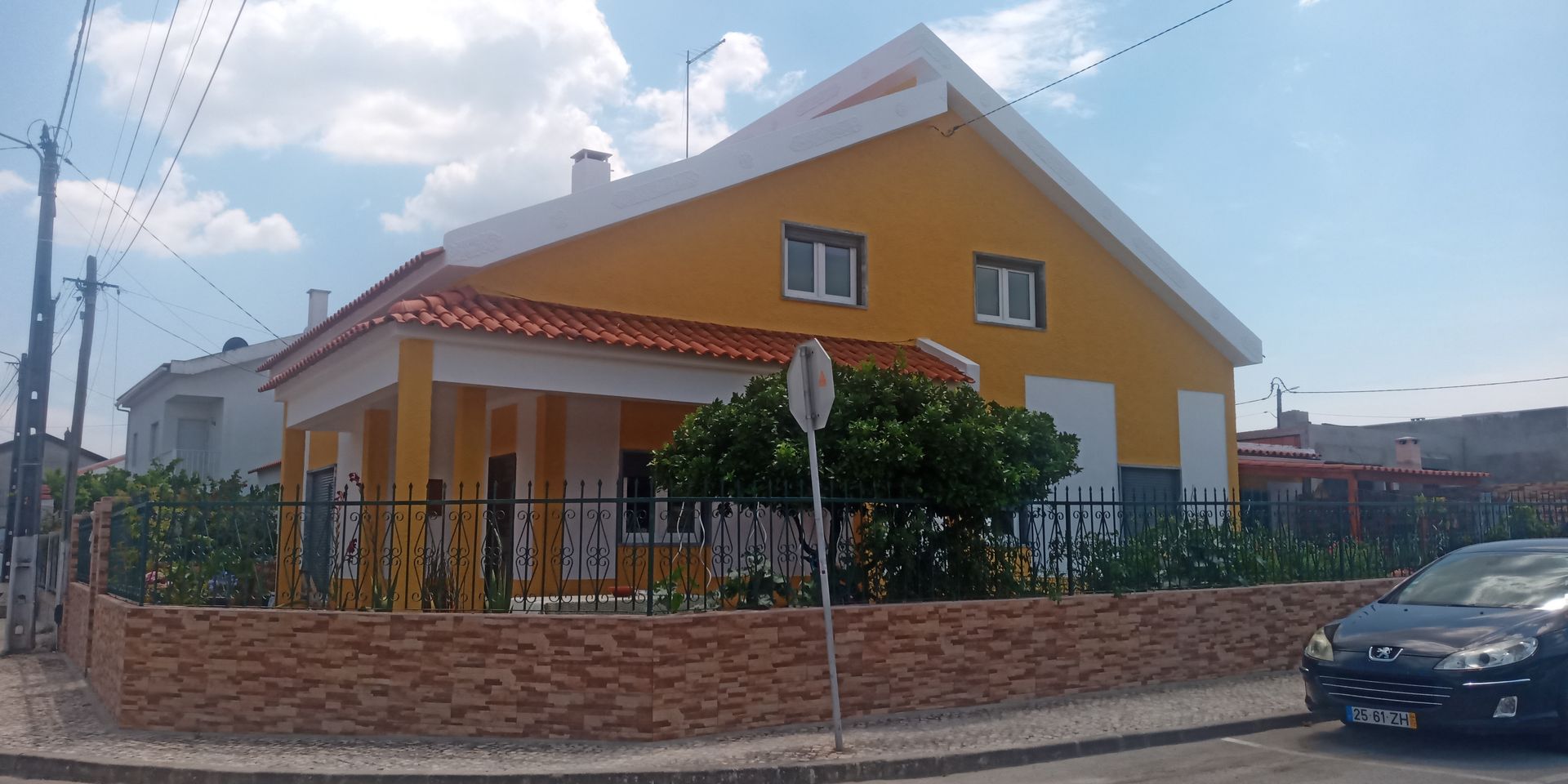 Casa amarela com detalhes brancos, telhado de telha vermelha e muro de tijolos, com um carro estacionado em frente.