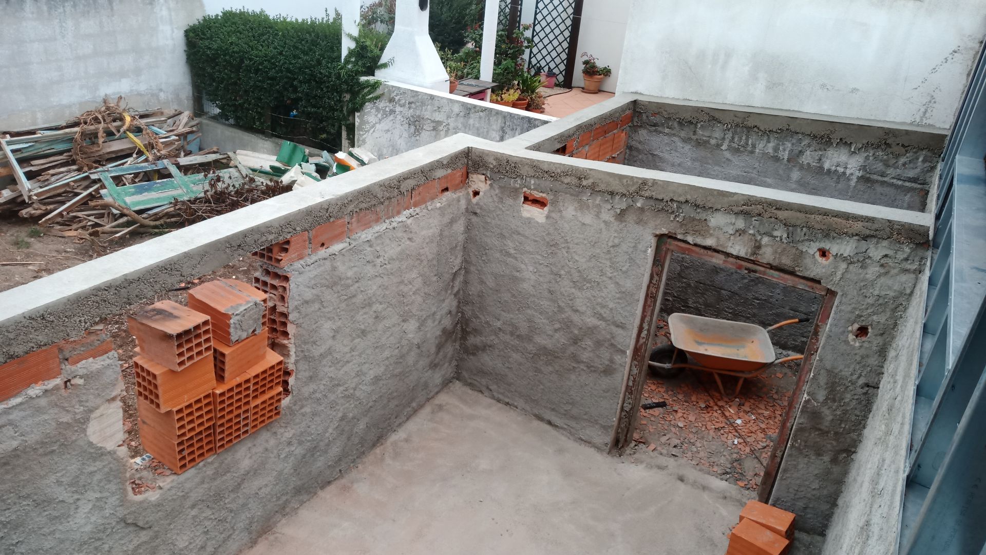 Canteiro de obras: paredes de concreto e tijolo de um prédio em construção, carrinho de mão na entrada, entulho ao fundo.