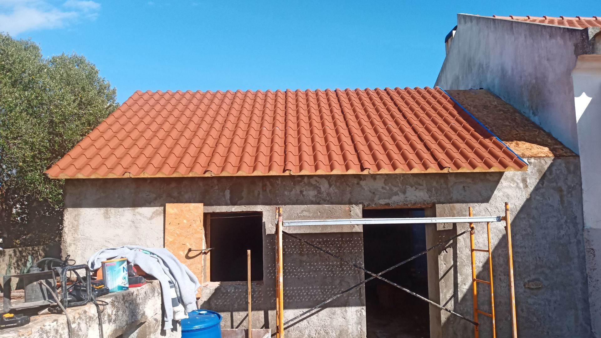 Um pequeno edifício em construção com telhado de telha vermelha, portas e janelas visíveis, andaimes e céu azul.