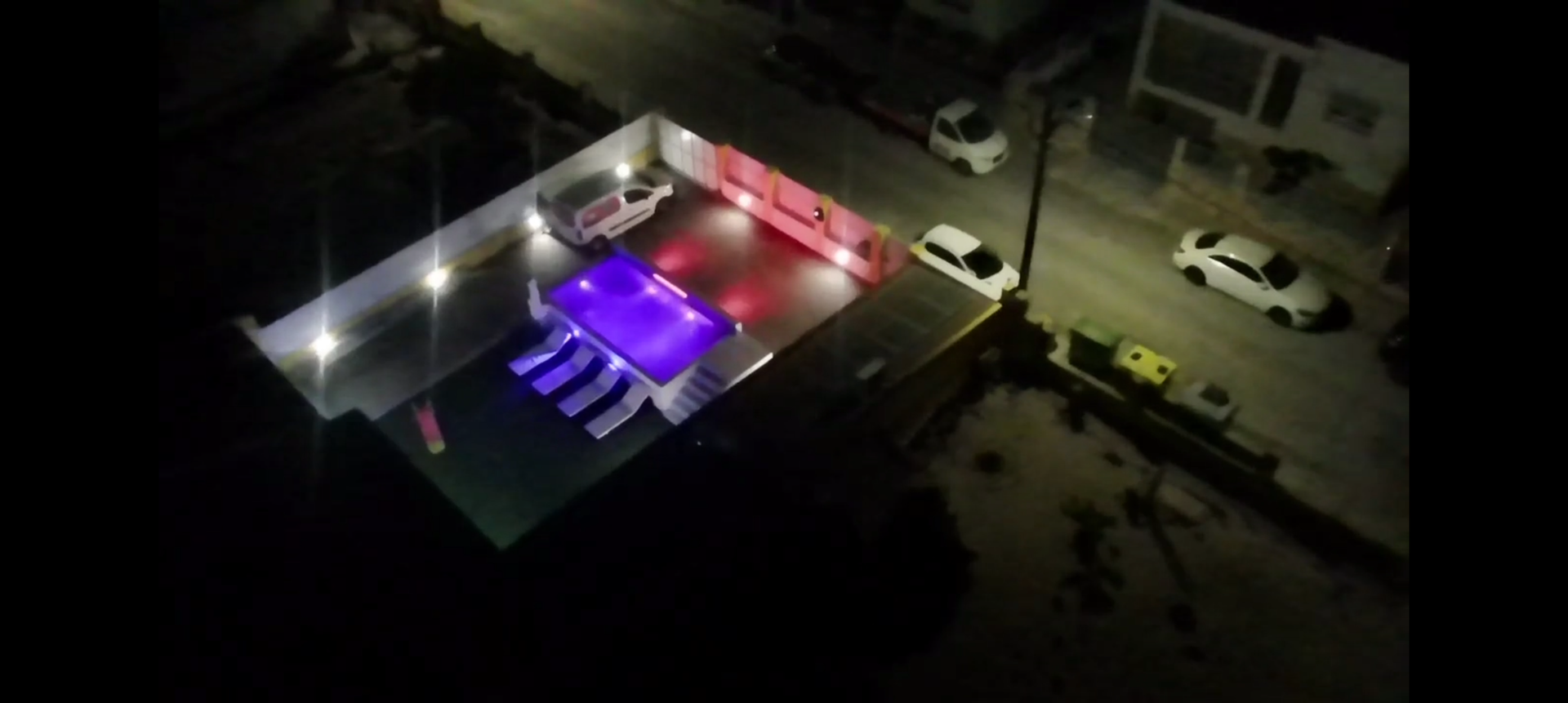 Vista aérea de uma piscina bem iluminada à noite, com carros brancos estacionados nas proximidades. Postes de luz iluminam a rua.