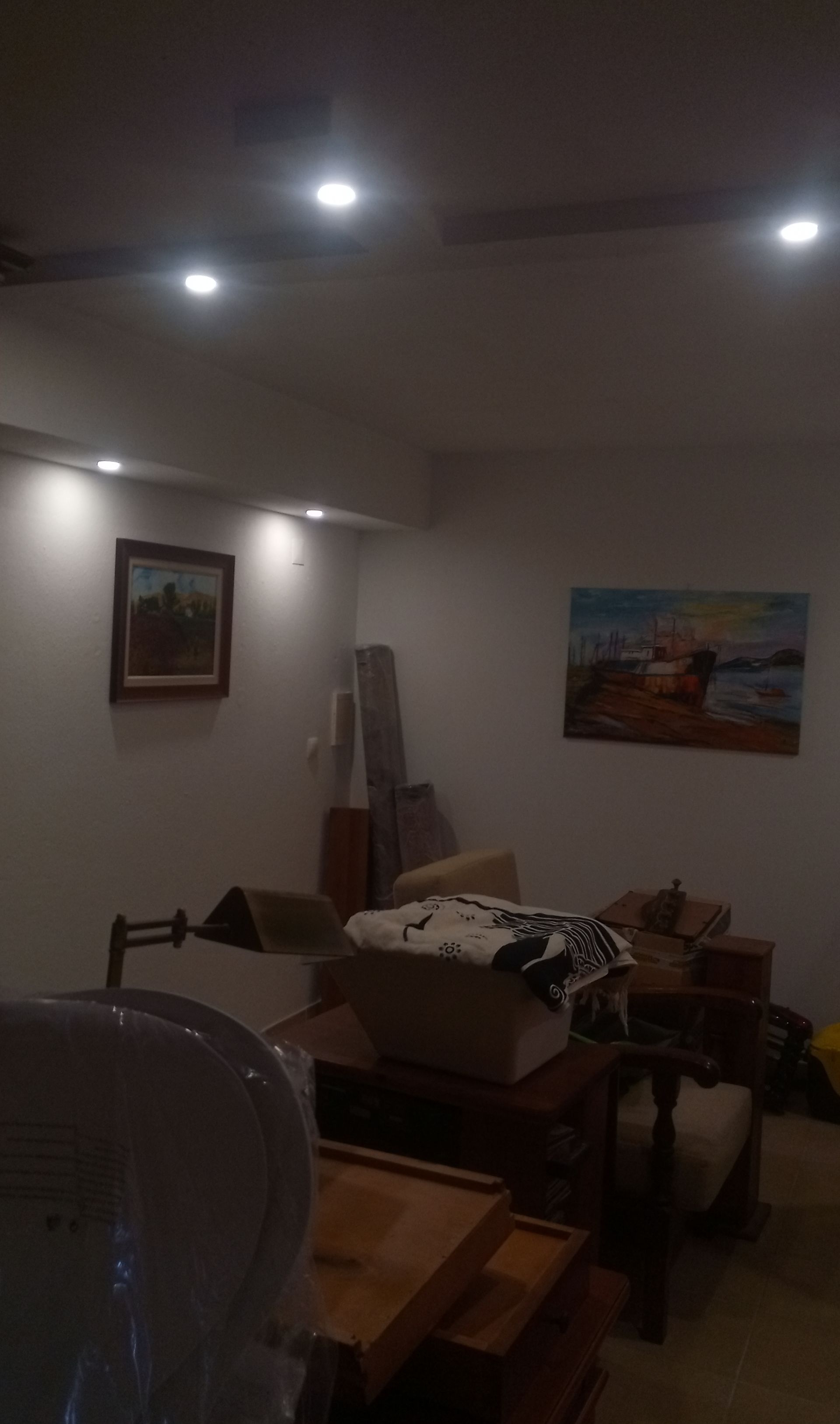 Um quarto com pouca luz, com móveis e quadros na parede. Luzes no teto iluminam a cena.