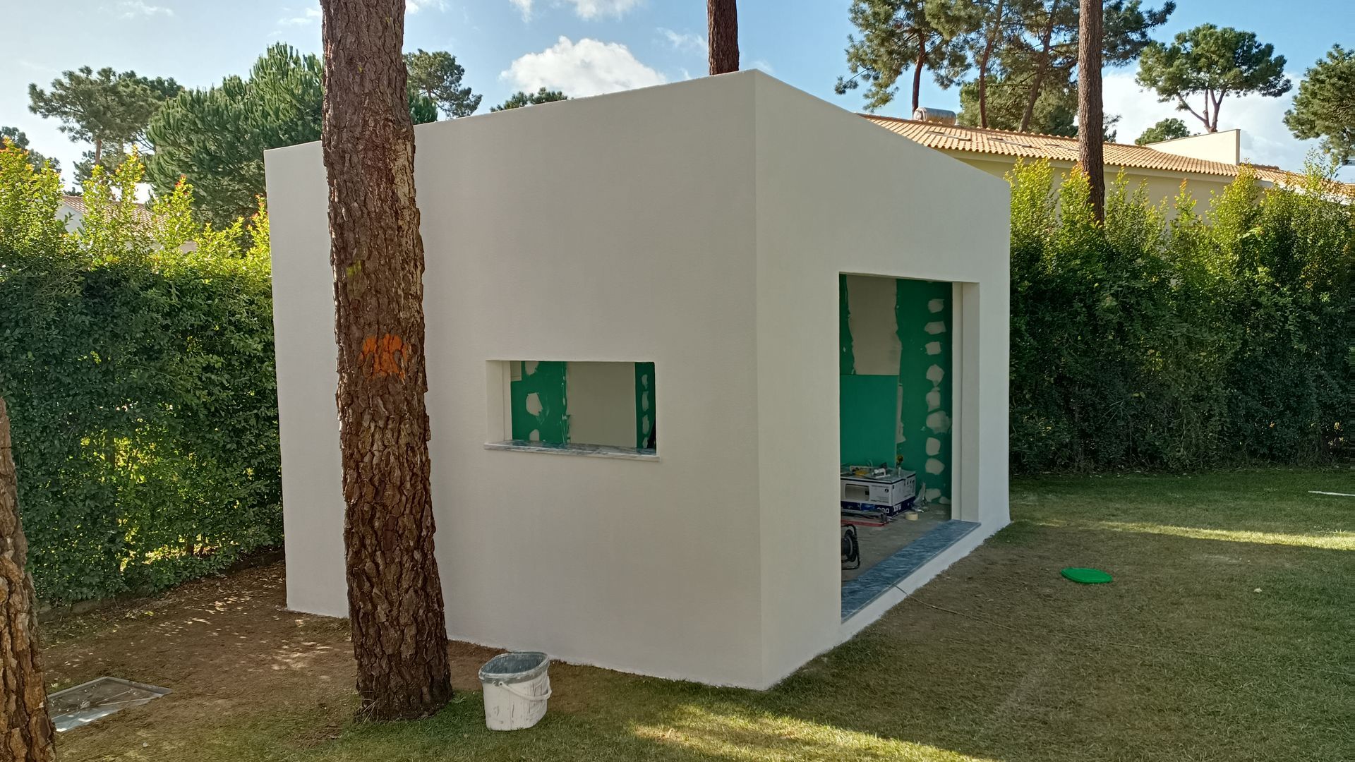Estrutura branca em forma de cubo com uma porta e janela abertas em um gramado verde, perto de uma árvore.