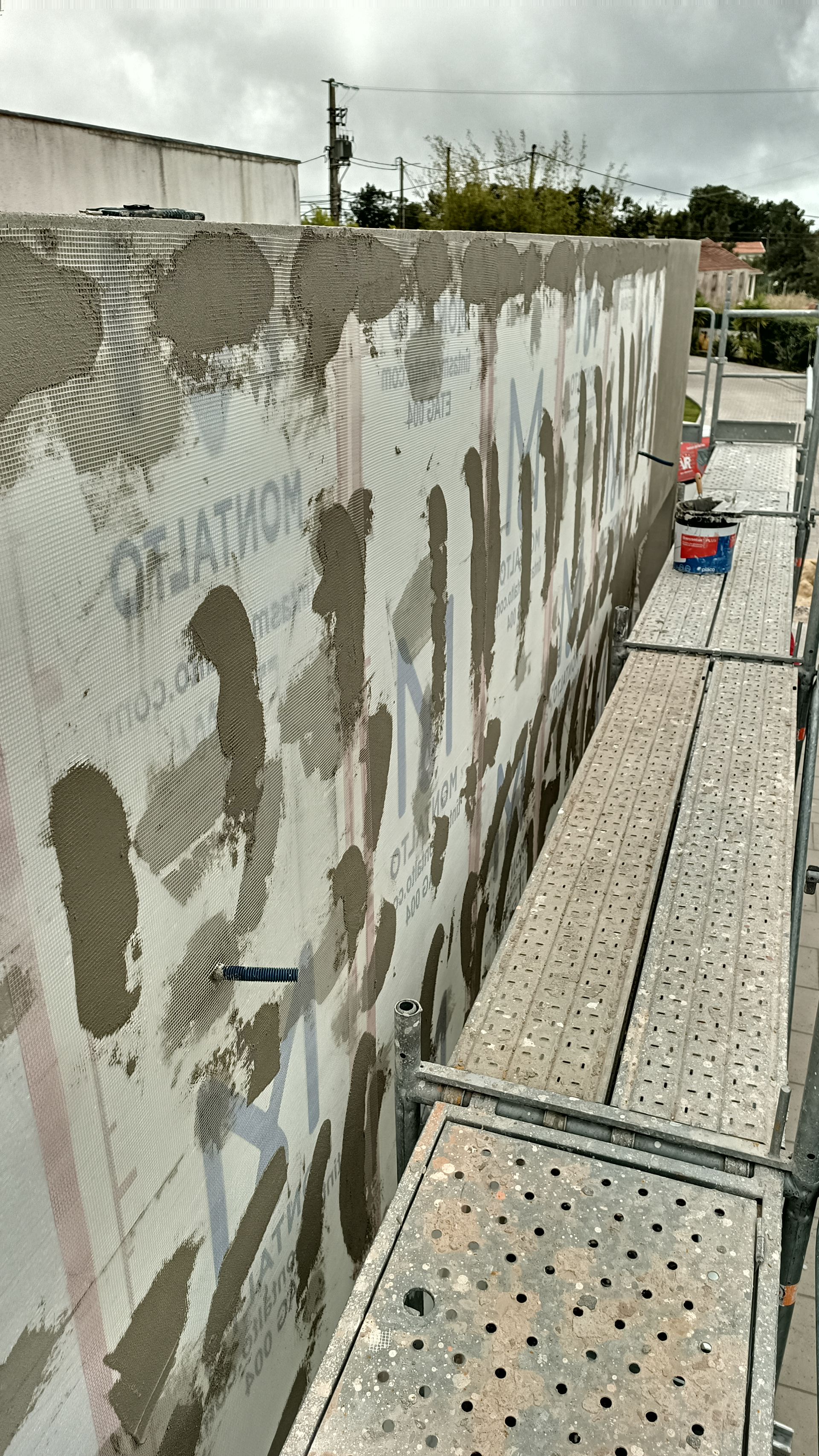 Andaimes montados ao longo de uma parede parcialmente revestida com gesso e tela metálica, em construção exterior.