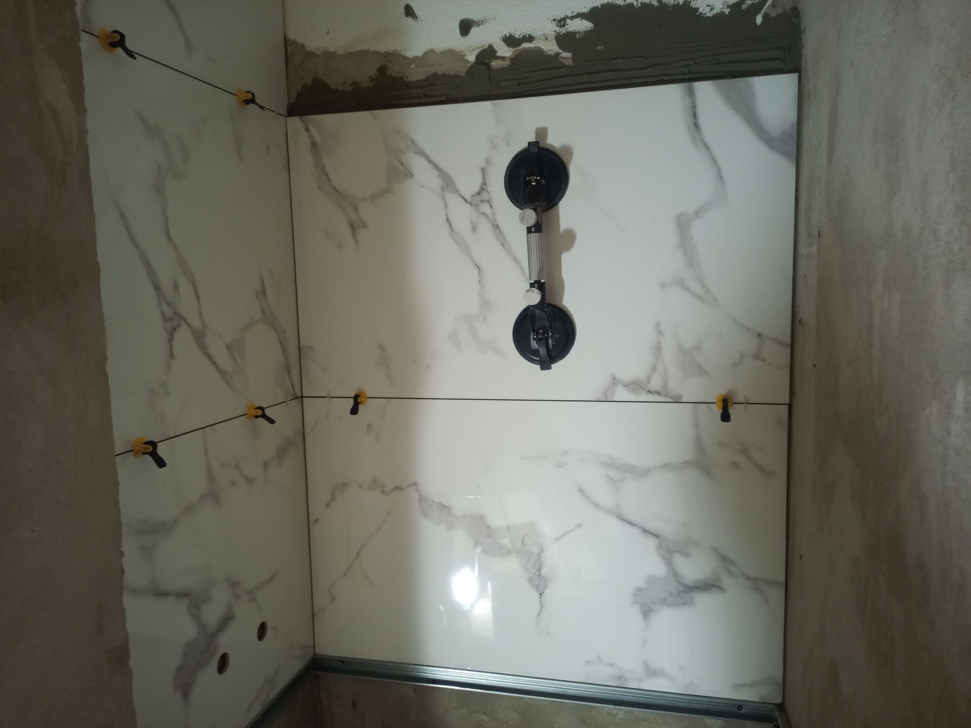 Parede do banheiro revestida com azulejos que imitam mármore e grampos de nivelamento, com as instalações hidráulicas visíveis.