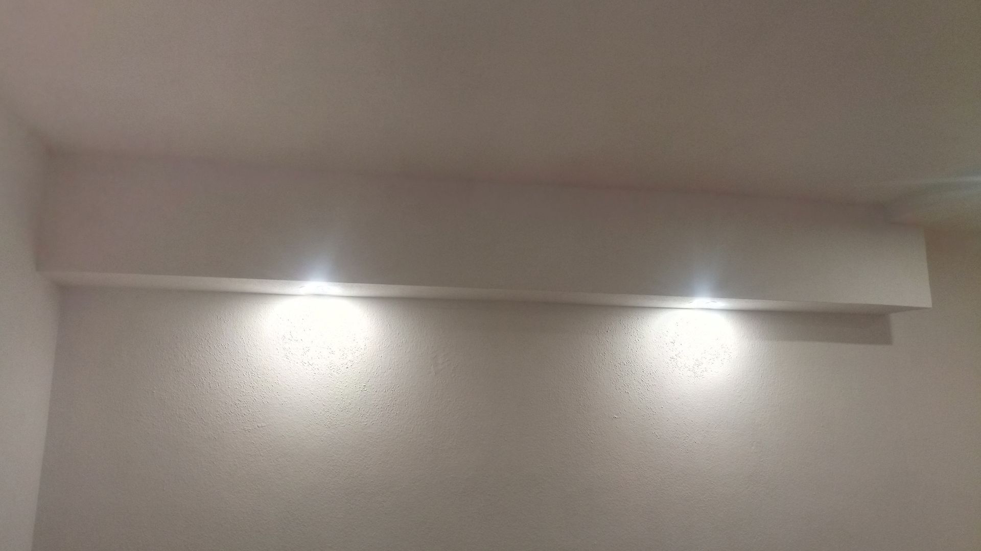 Parede branca com uma prateleira e duas luzes embutidas iluminando a parede.