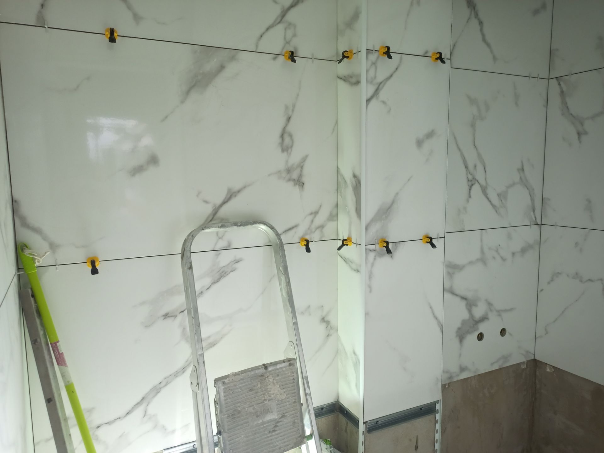 Azulejos brancos com estampa de mármore sendo instalados na parede de um banheiro, utilizando grampos niveladores. Uma escada e um nível estão presentes.