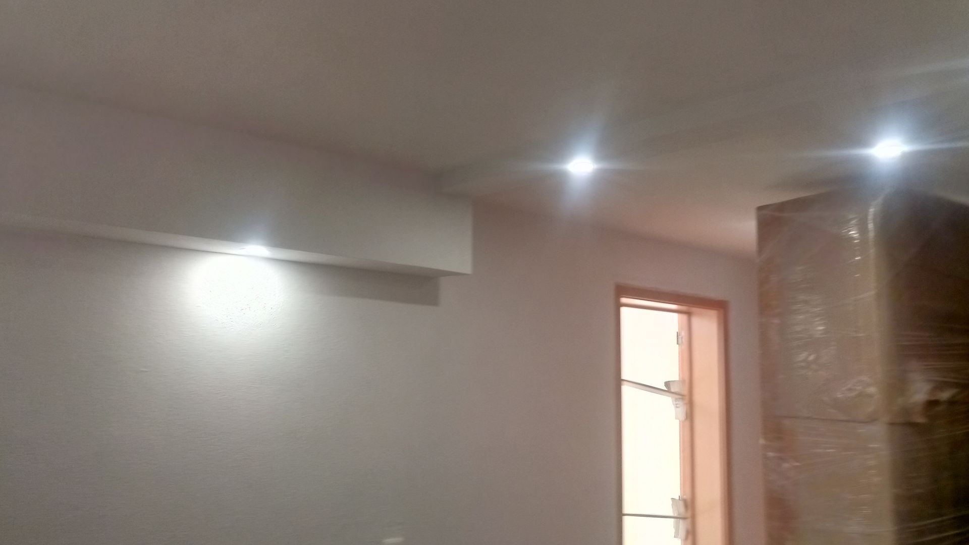 Quarto branco com luzes embutidas iluminando o teto. Uma janela e móveis cobertos são visíveis.