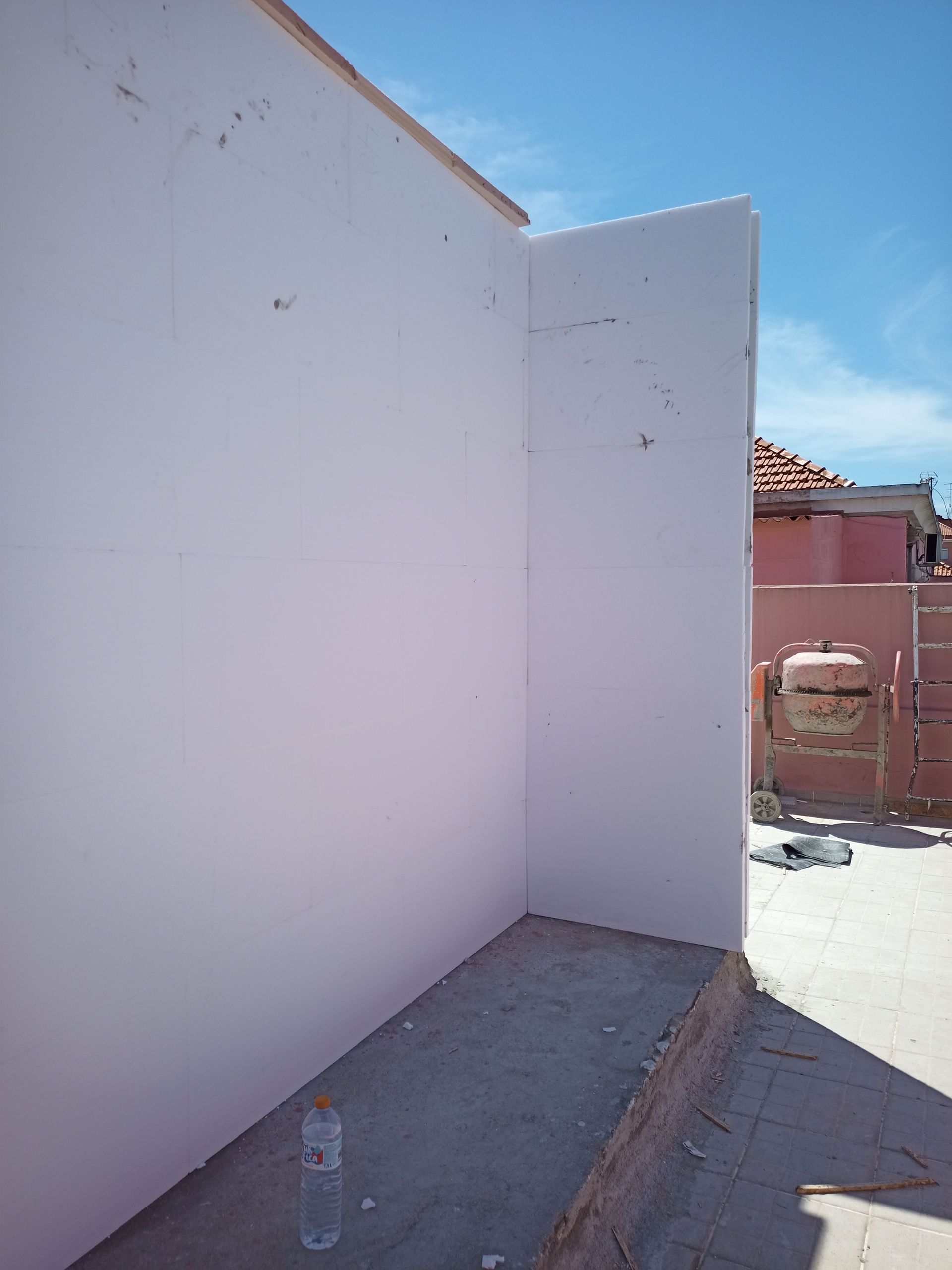 Paredes brancas nos cantos, base de concreto, garrafa no chão, céu azul ao fundo.