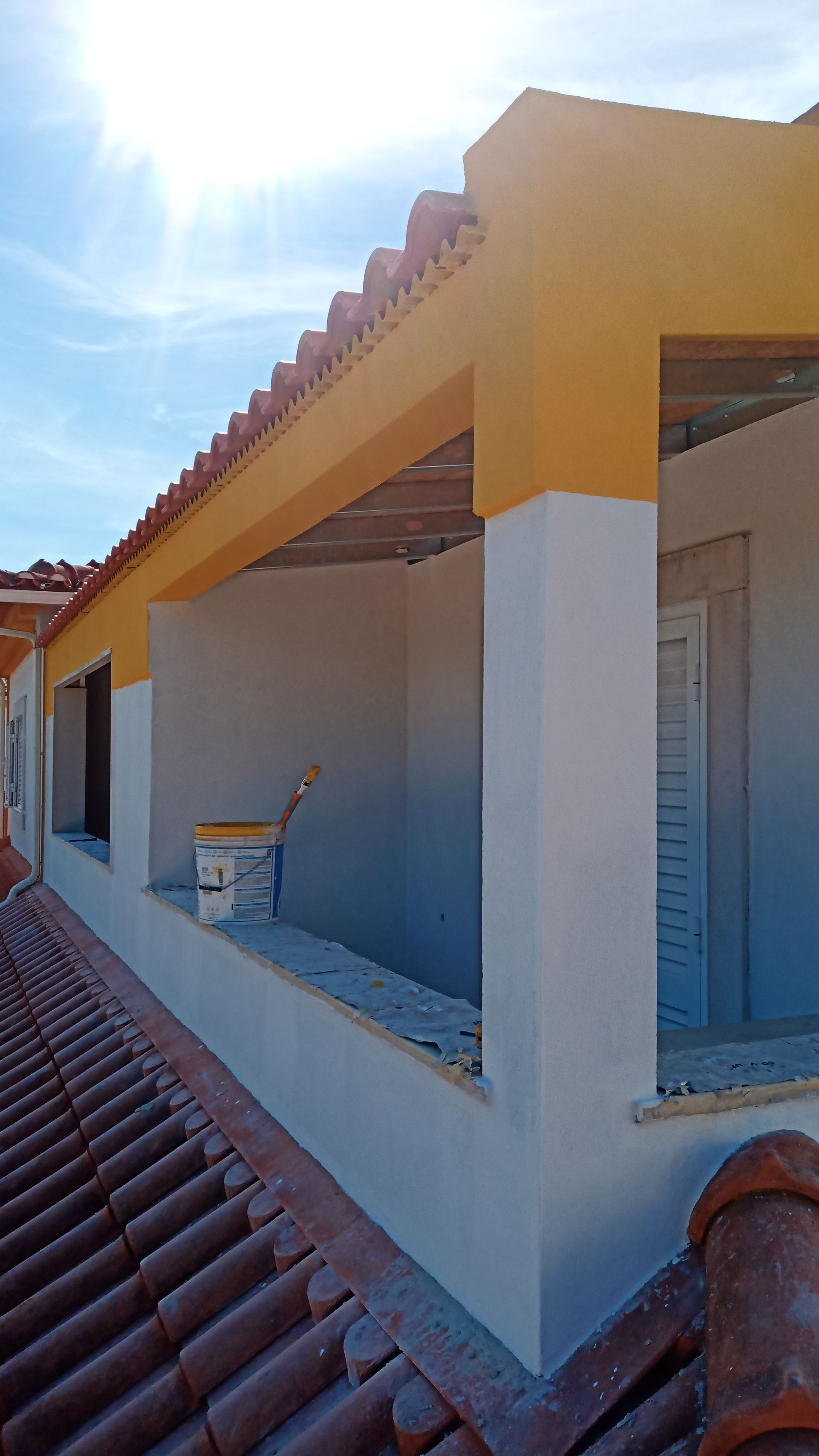 Vista exterior de um edifício com paredes brancas, detalhes em amarelo e telhado de telha vermelha, parcialmente em construção.