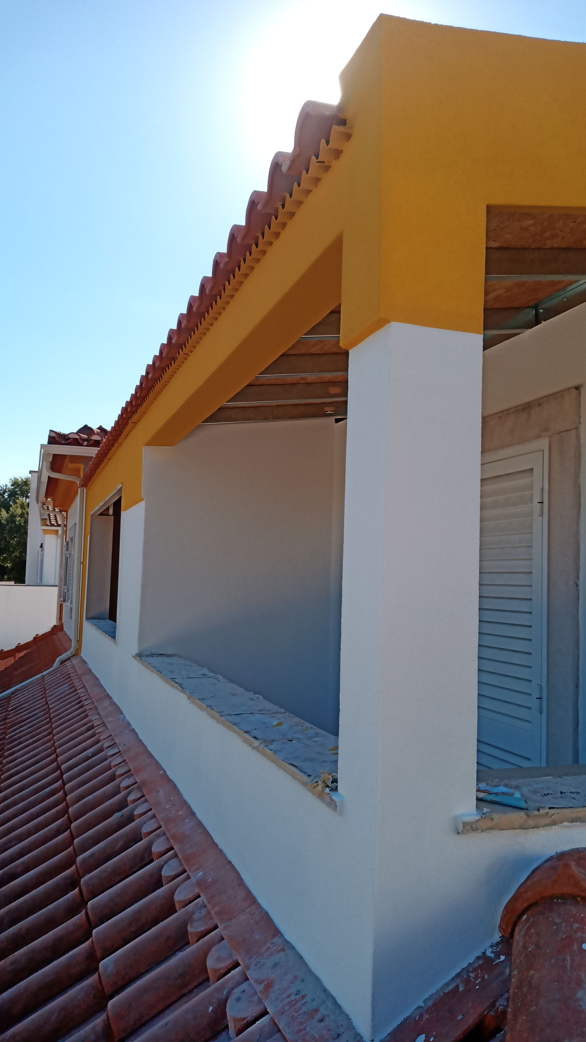 Telhado de telhas laranja com beiral pintado de amarelo, pilares e paredes brancas, céu azul.