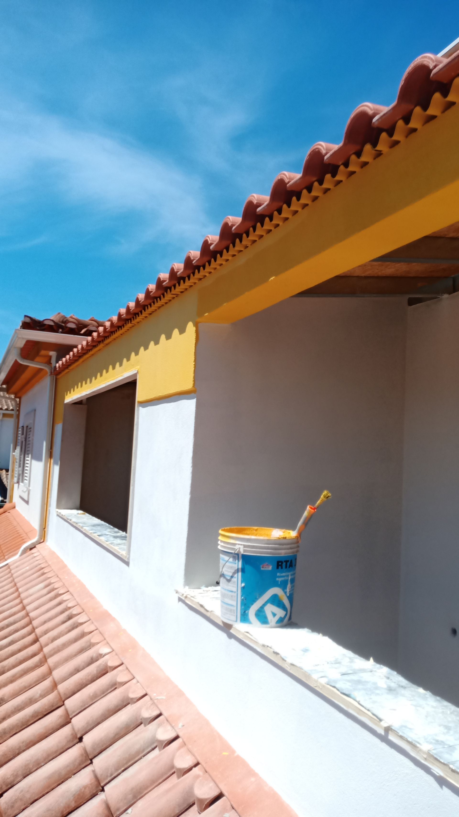 Exterior de um edifício em construção, com telhado amarelo, telhas de terracota e um balde de tinta.