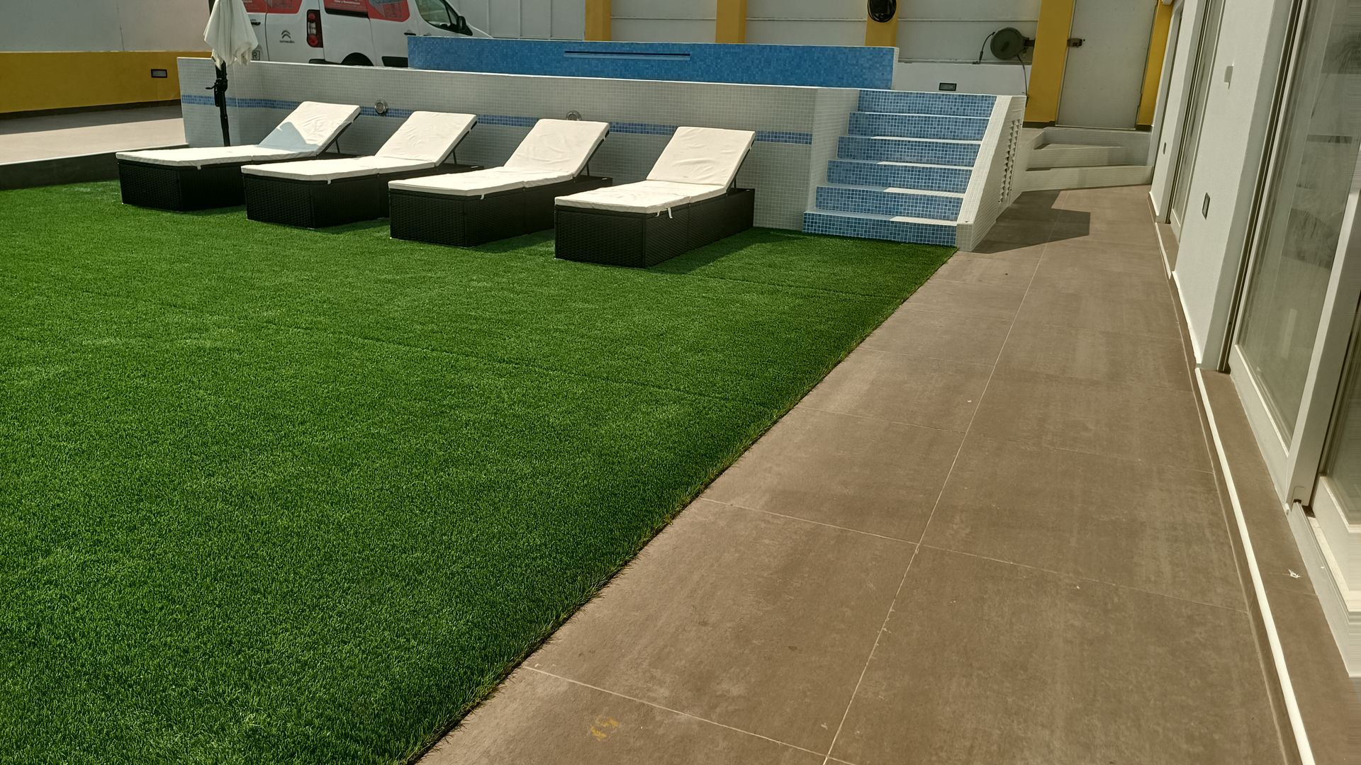 Cadeiras de descanso em grama artificial verde ao lado de uma passarela marrom, perto de um muro e de um prédio.