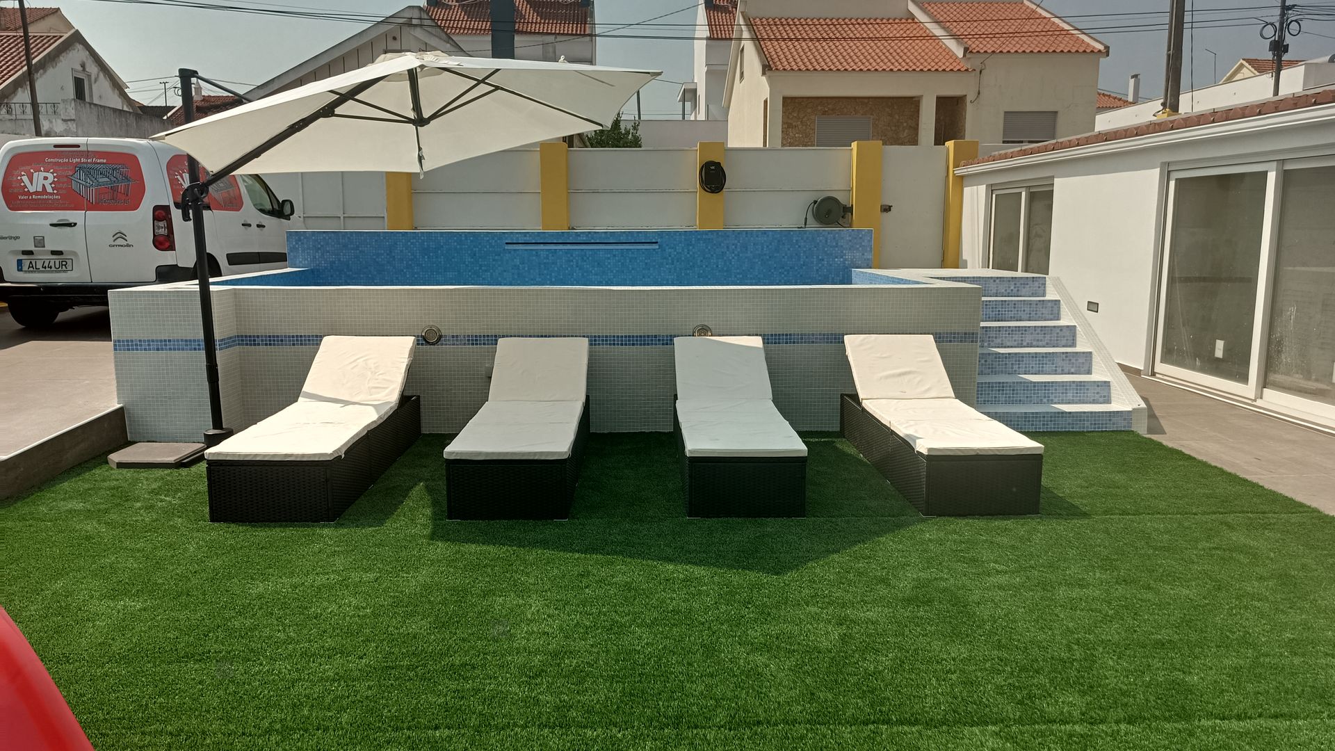 Área da piscina com espreguiçadeiras, guarda-sol e grama sintética.